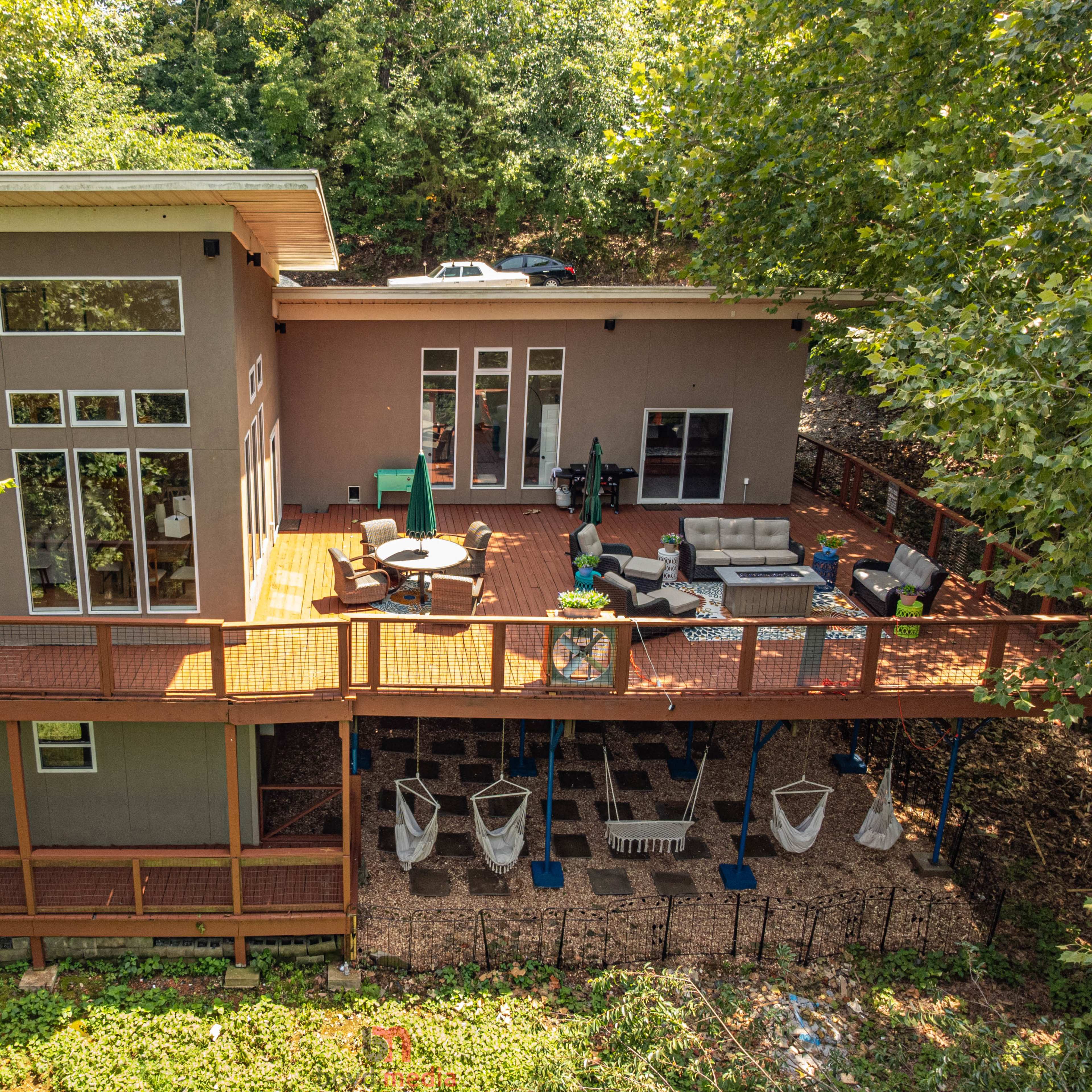 A modern house with a large deck surrounded by trees, featuring outdoor furniture and a garden area below.