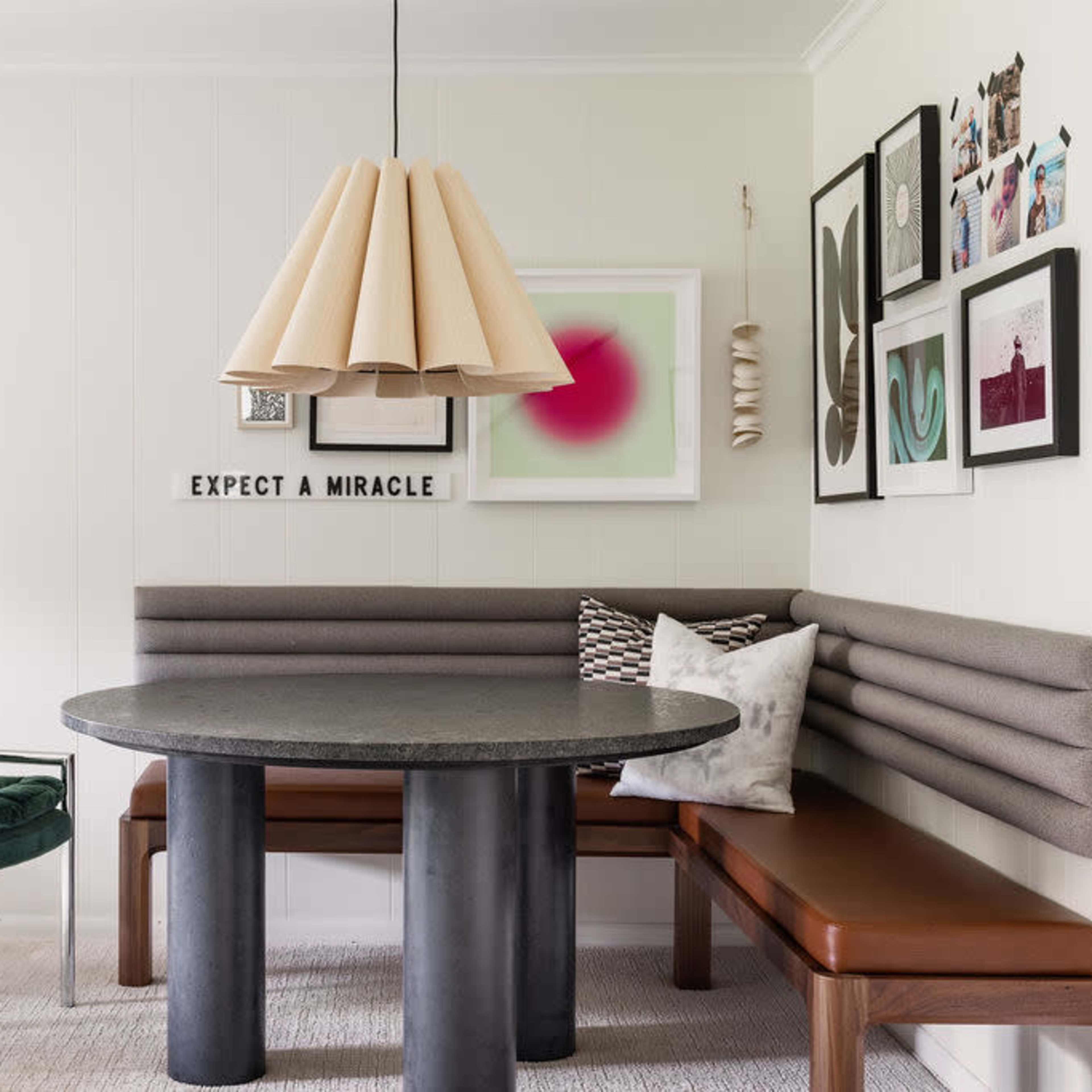 A modern dining area with a round table, a bench with backrest seating, and a green chair, adorned with several framed artworks on the wall.