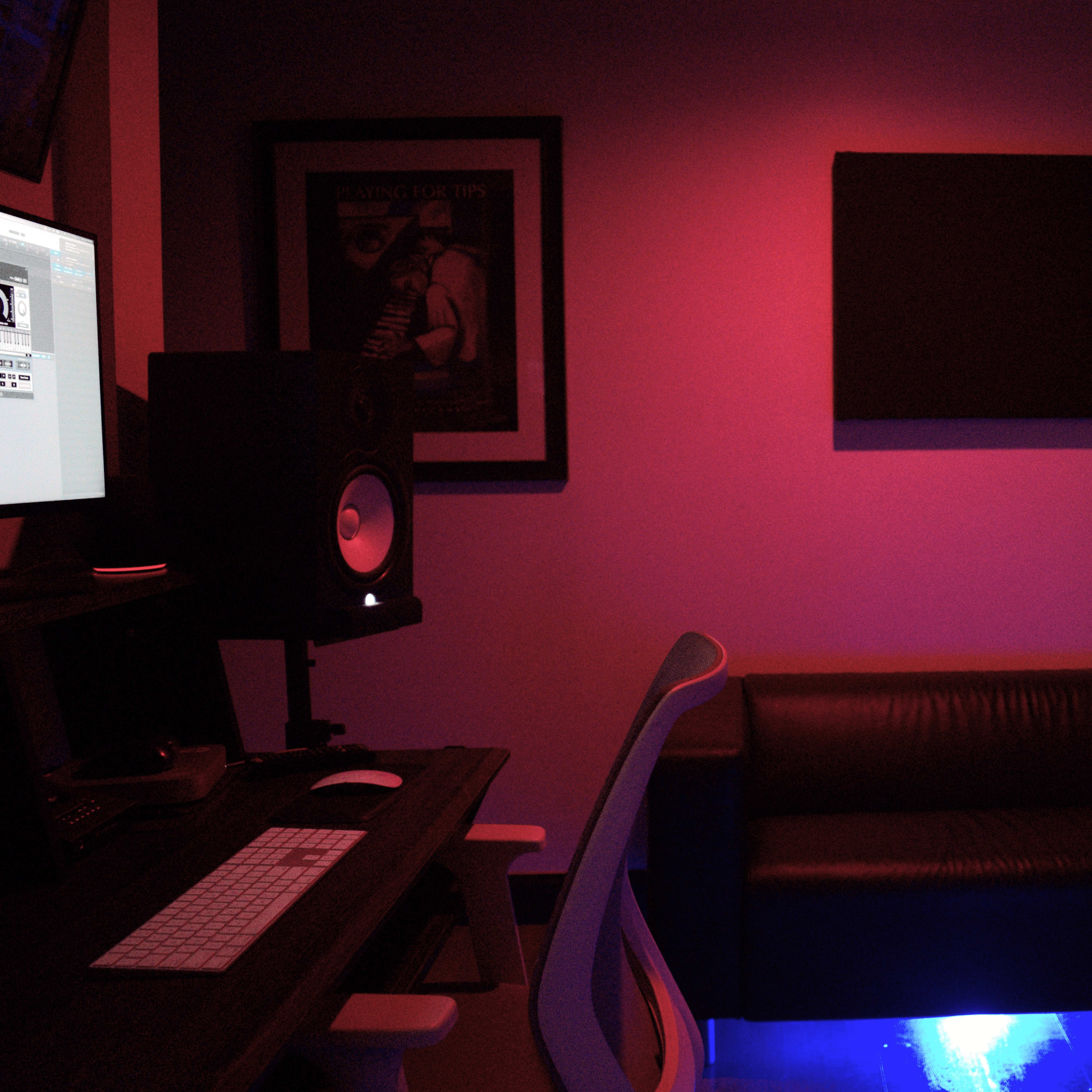 A music studio features a darkened space with a computer desk, an audio speaker, and a black couch illuminated by blue lighting.