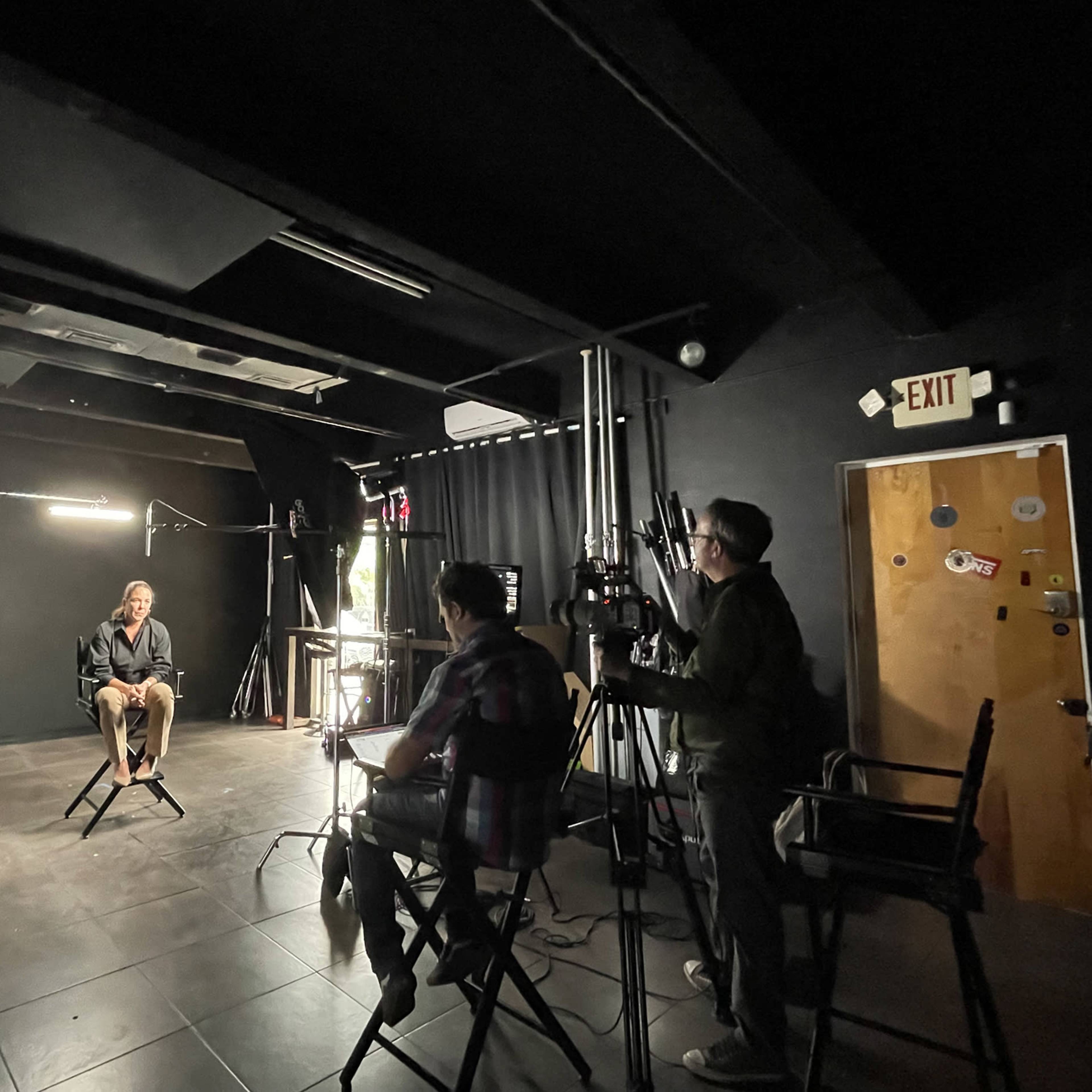 A man is sitting in a chair under bright lights while two individuals operate cameras in a dark studio setting.