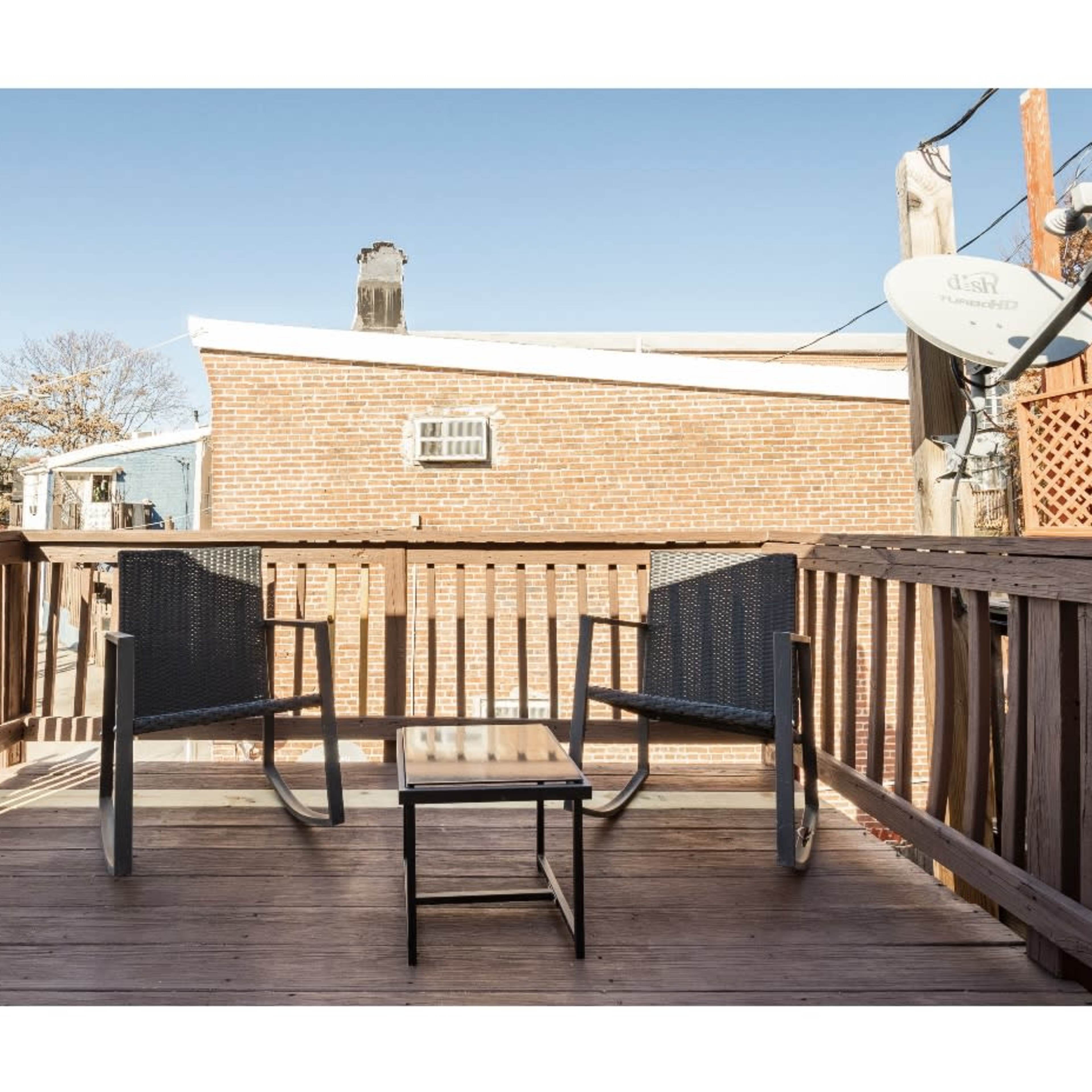 A small wooden deck with two chairs and a table, surrounded by brick buildings and a clear sky.