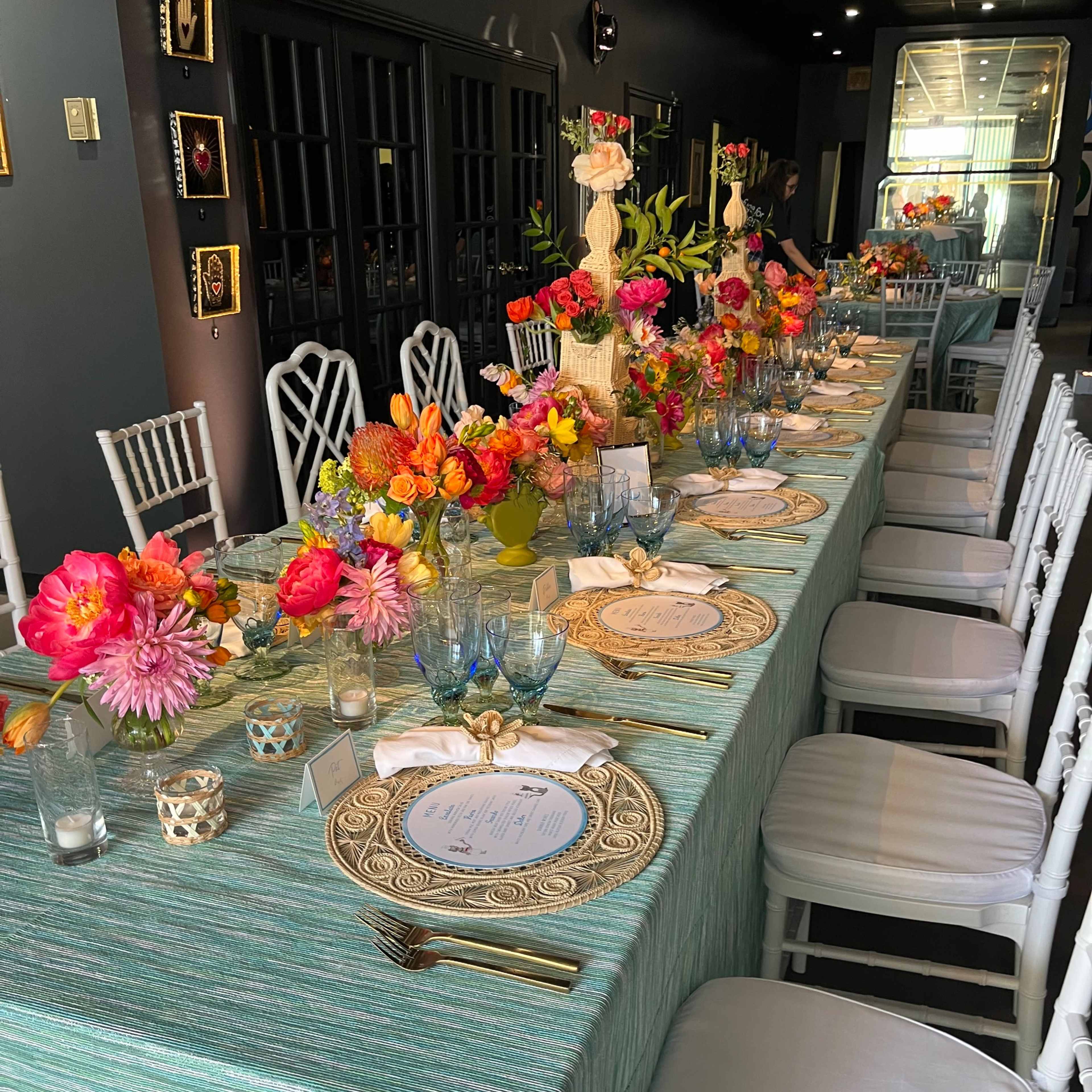 A long dining table is decorated with vibrant floral centerpieces, elegant tableware, and a light blue tablecloth in a well-lit indoor setting.