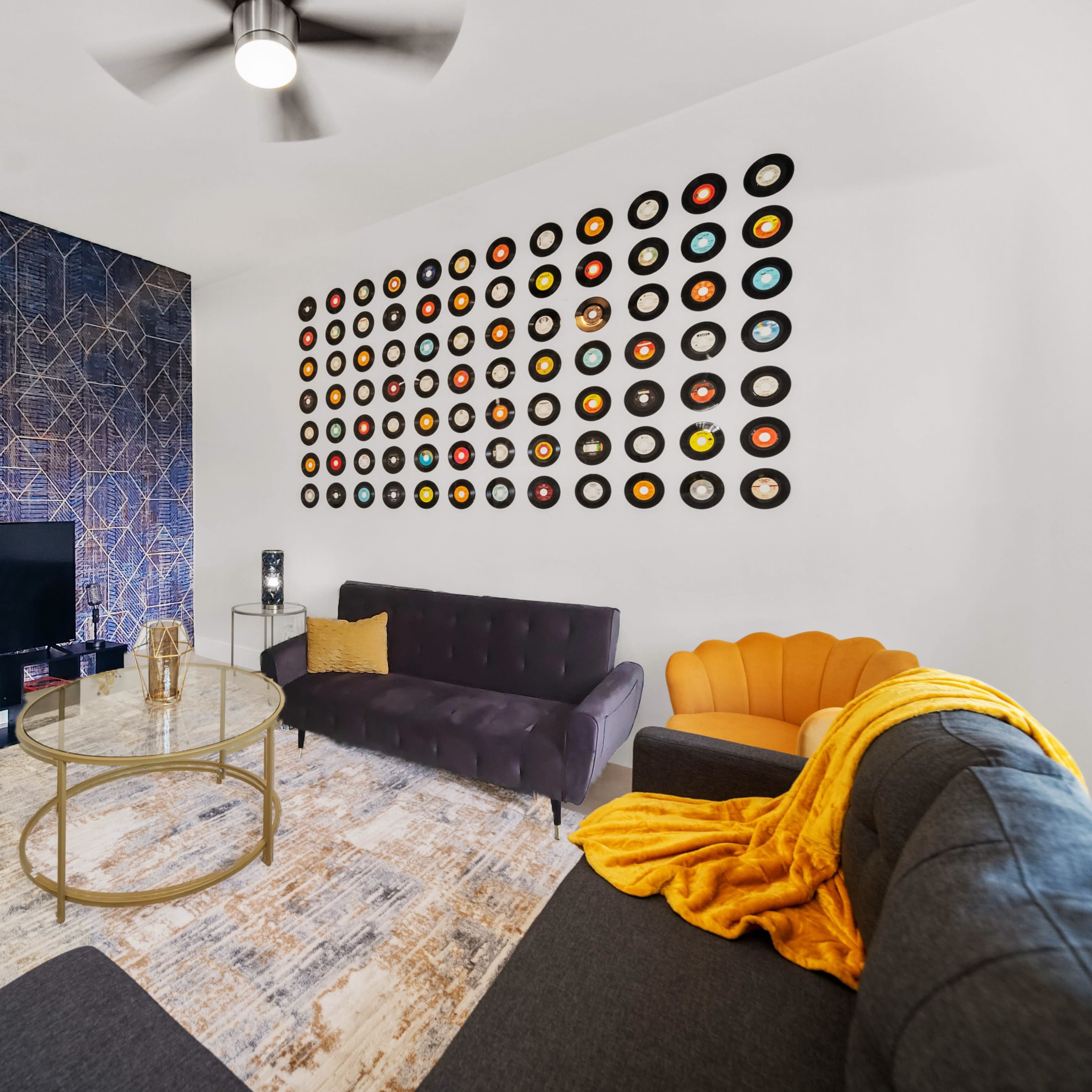 A modern living room features a patterned wall, a television mounted above a low table, a dark sectional sofa, an orange chair, and a collection of vinyl records arranged on the wall.