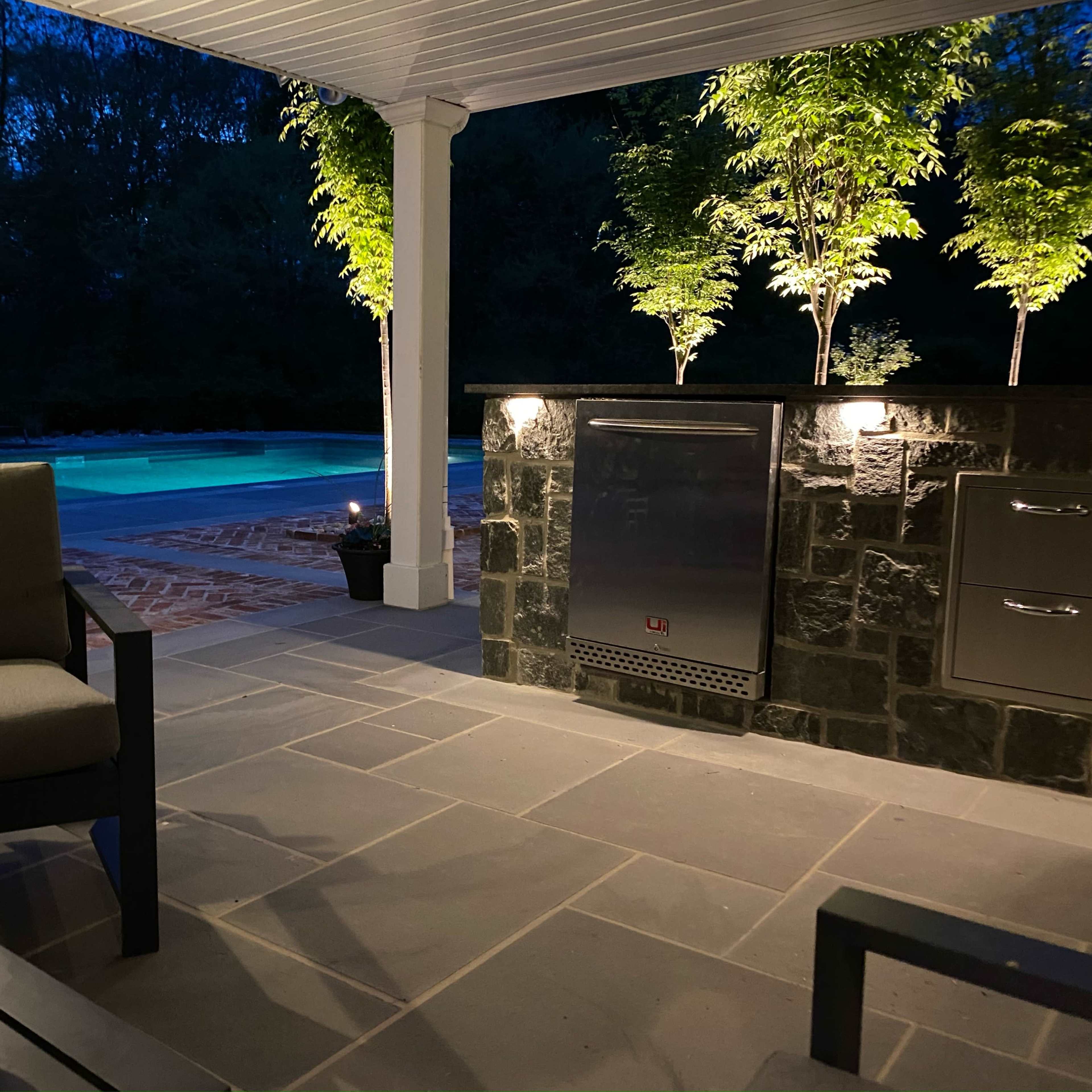 A calm outdoor seating area with modern furniture, a stone countertop, and a swimming pool illuminated in the background under the evening sky.