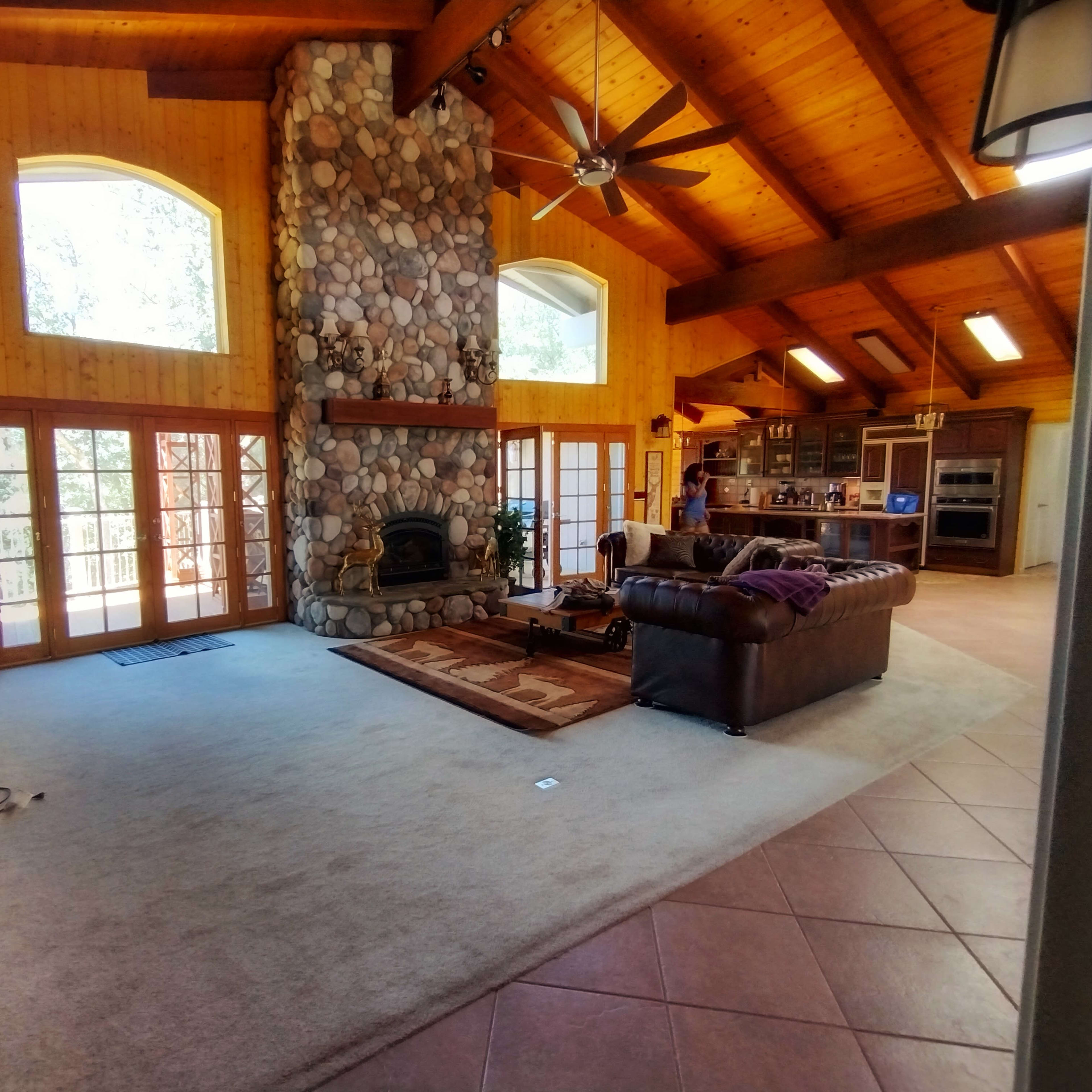 A spacious room features a stone fireplace, wooden beams, and large windows, with a couch and kitchen area visible in the background.