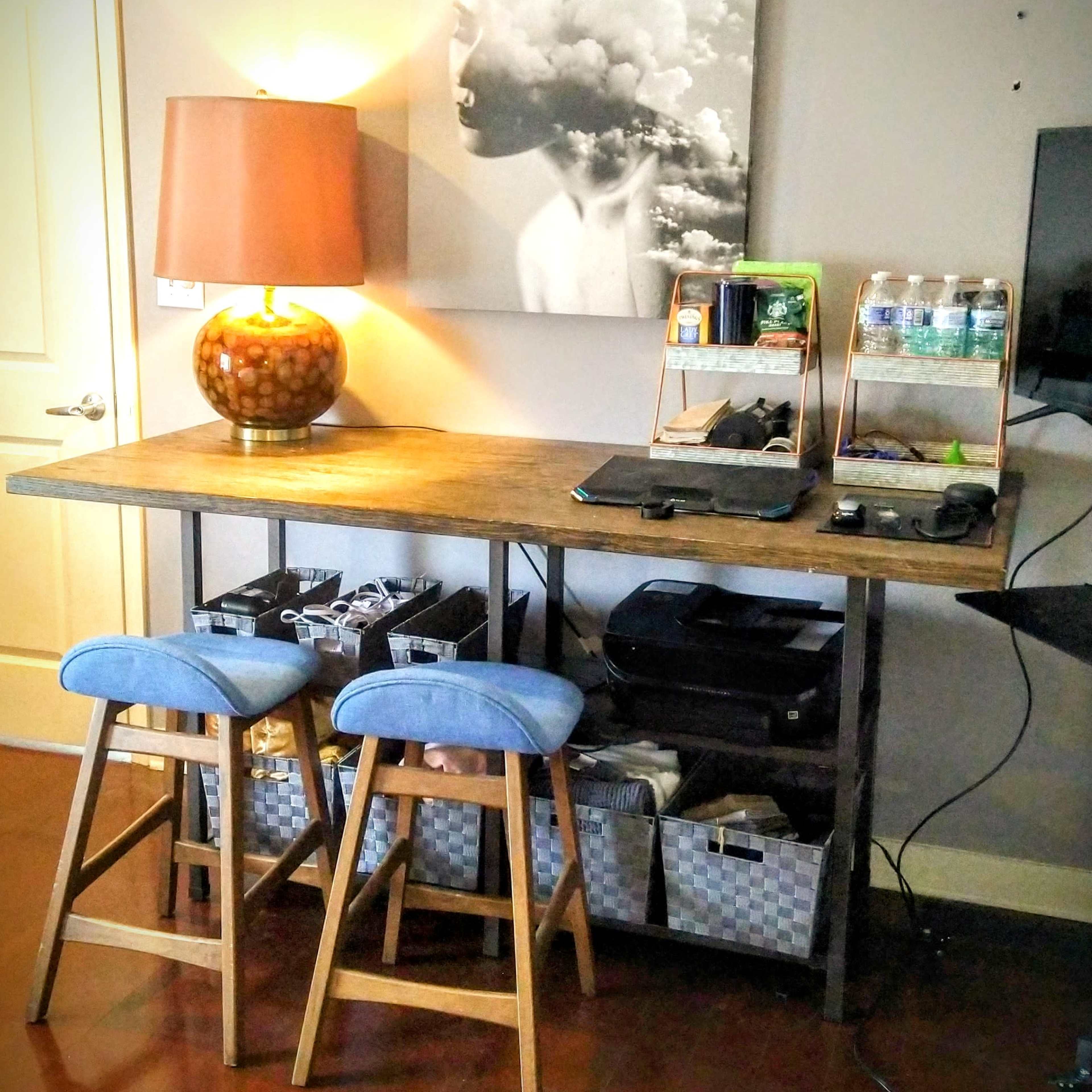 A wooden table with two blue stools is positioned against a wall featuring a large black-and-white artwork, with a lamp and organizational shelves on top.