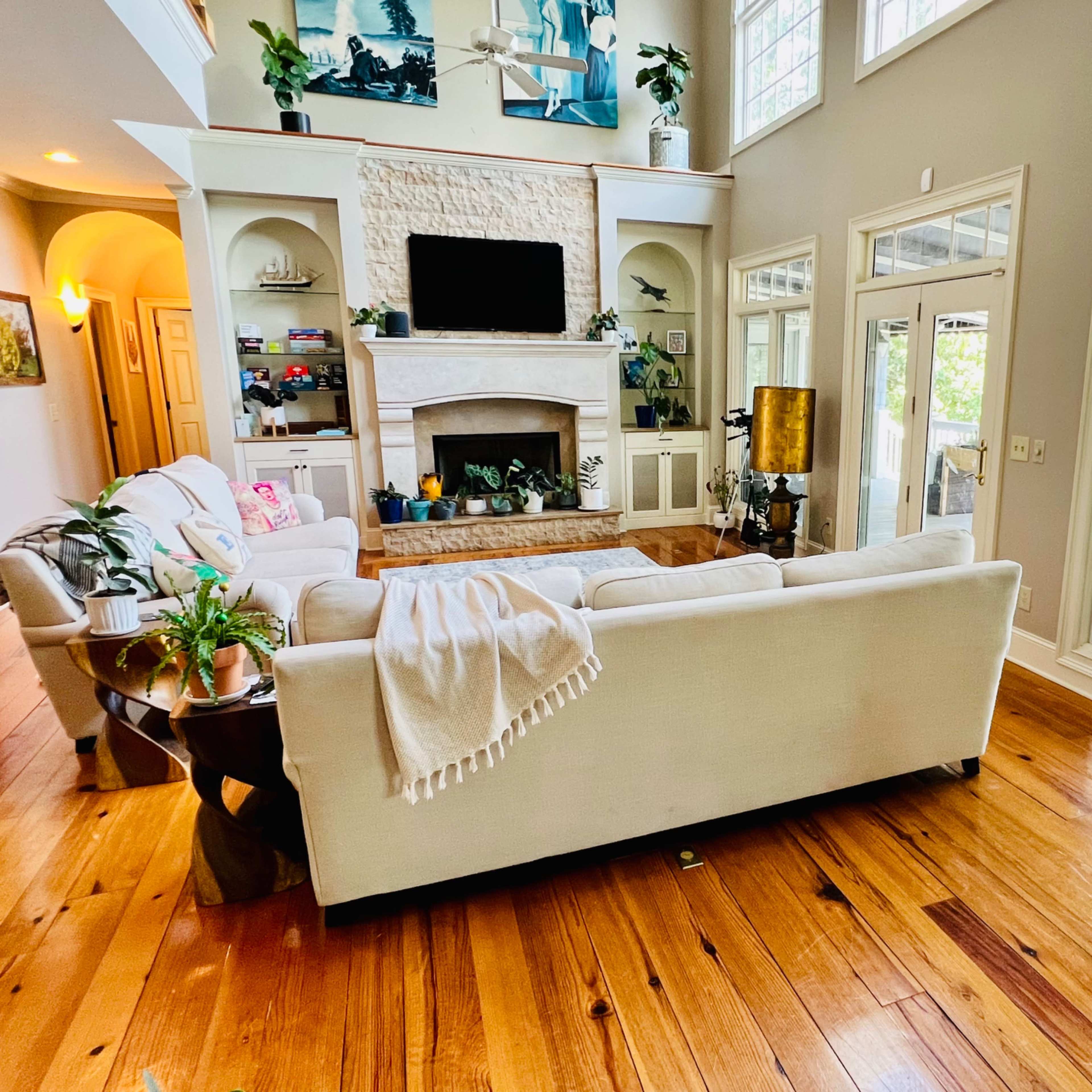 A spacious living room features a large white sofa, wooden flooring, and a stone fireplace, with windows that overlook a deck.
