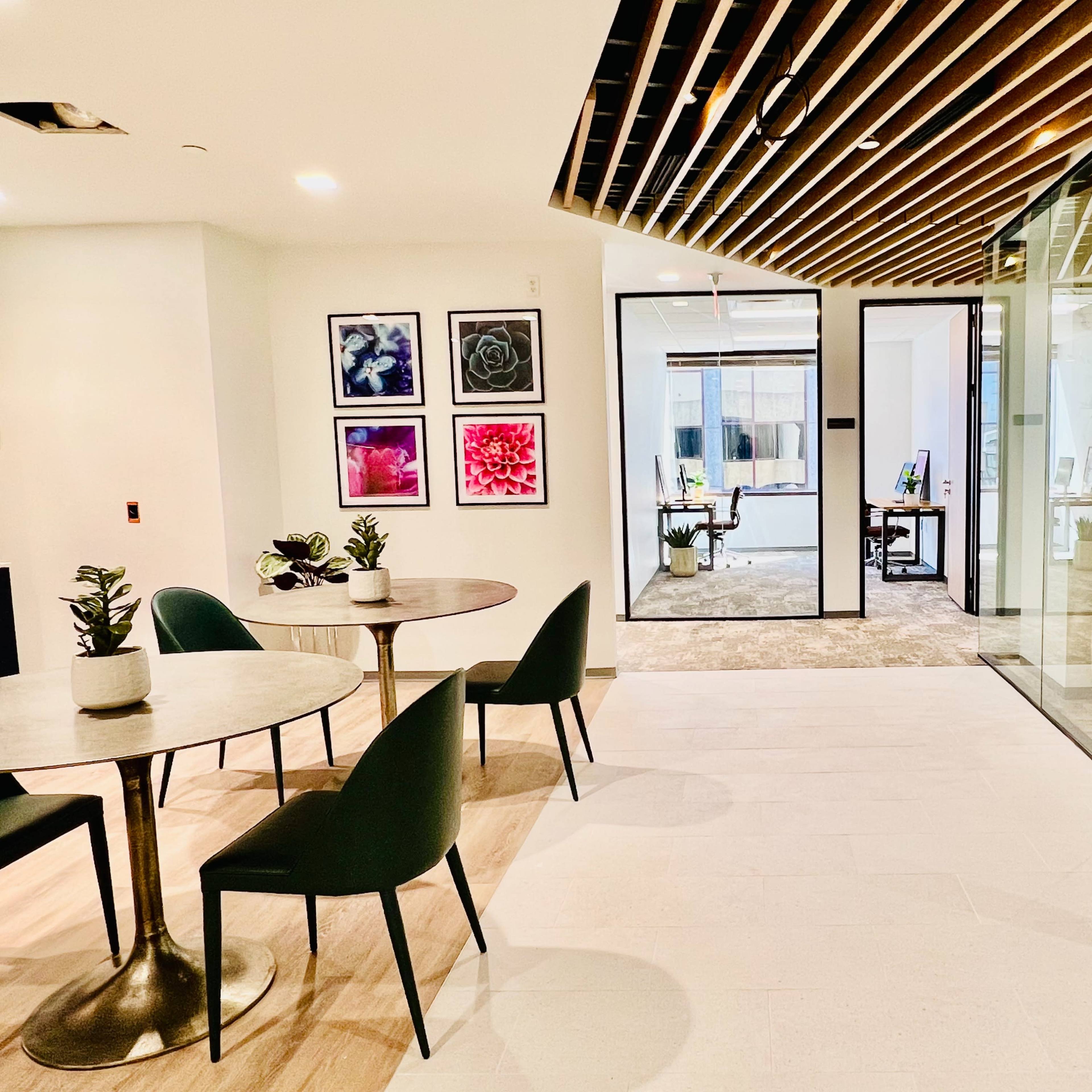 A modern office space featuring two round tables with green chairs, framed floral artwork on the walls, and a glass partition leading to another room.