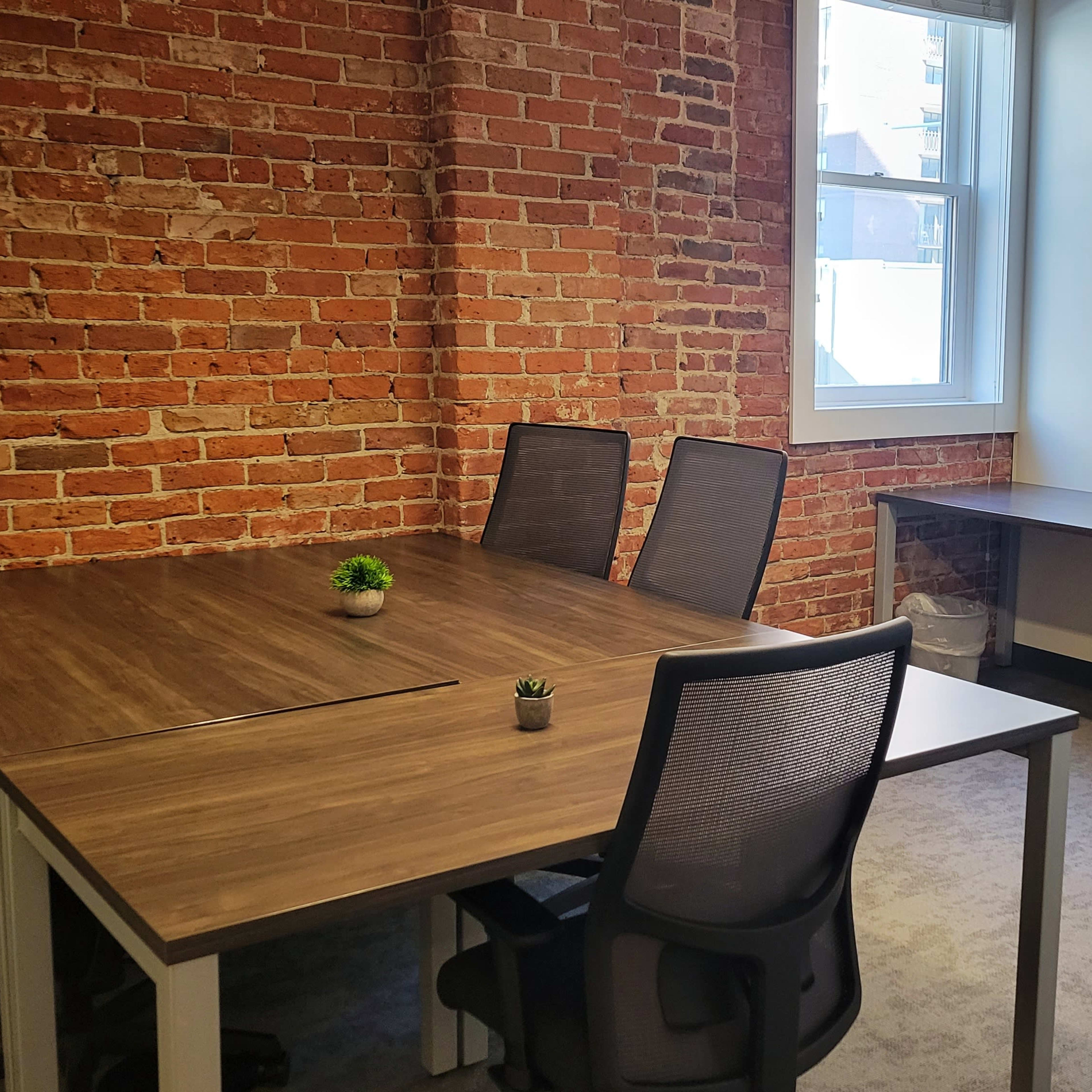 A modern office space features a wooden table surrounded by four chairs, with a brick wall in the background and a small plant on the table.