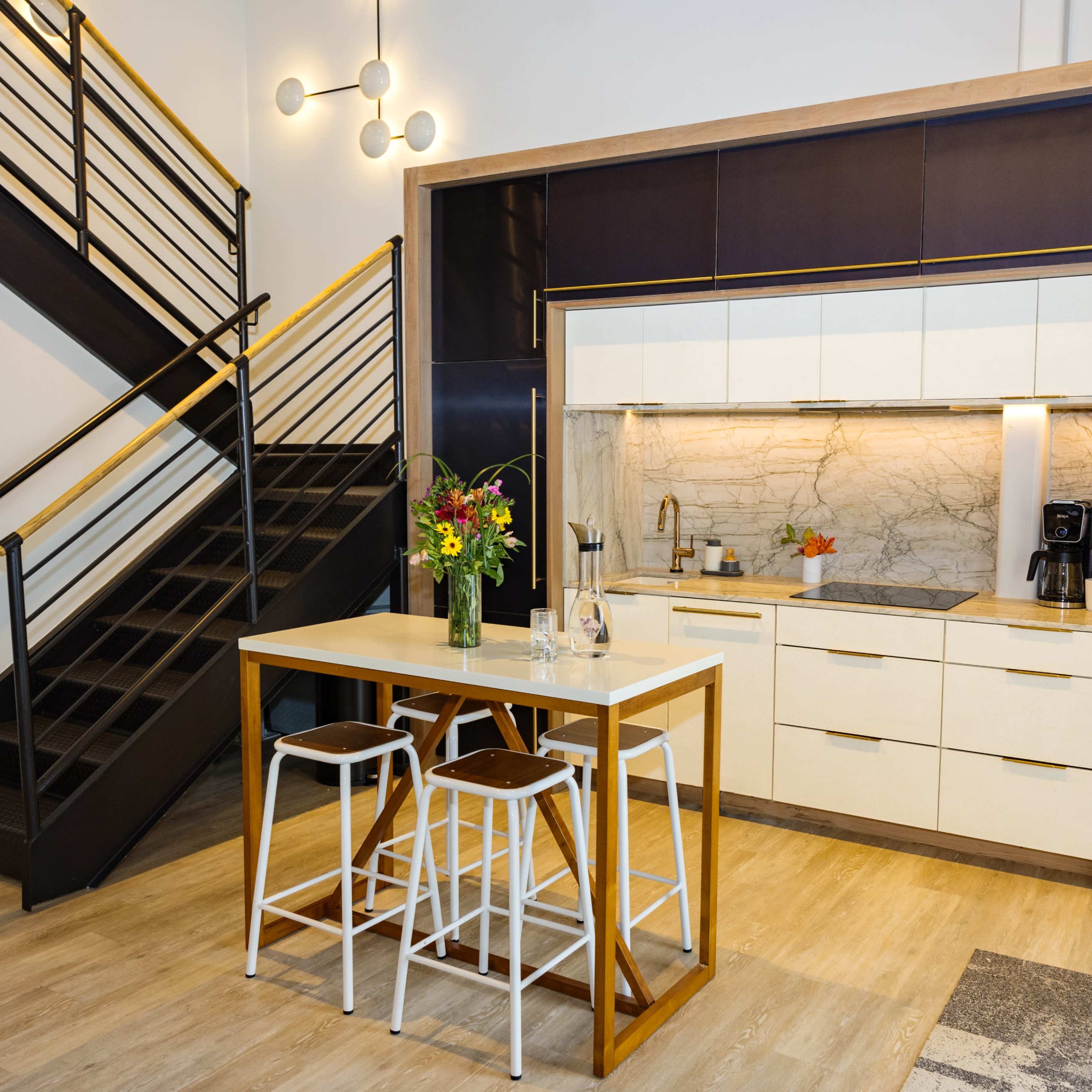 A modern kitchen features a central island with bar stools, sleek cabinetry, and a staircase in the background.