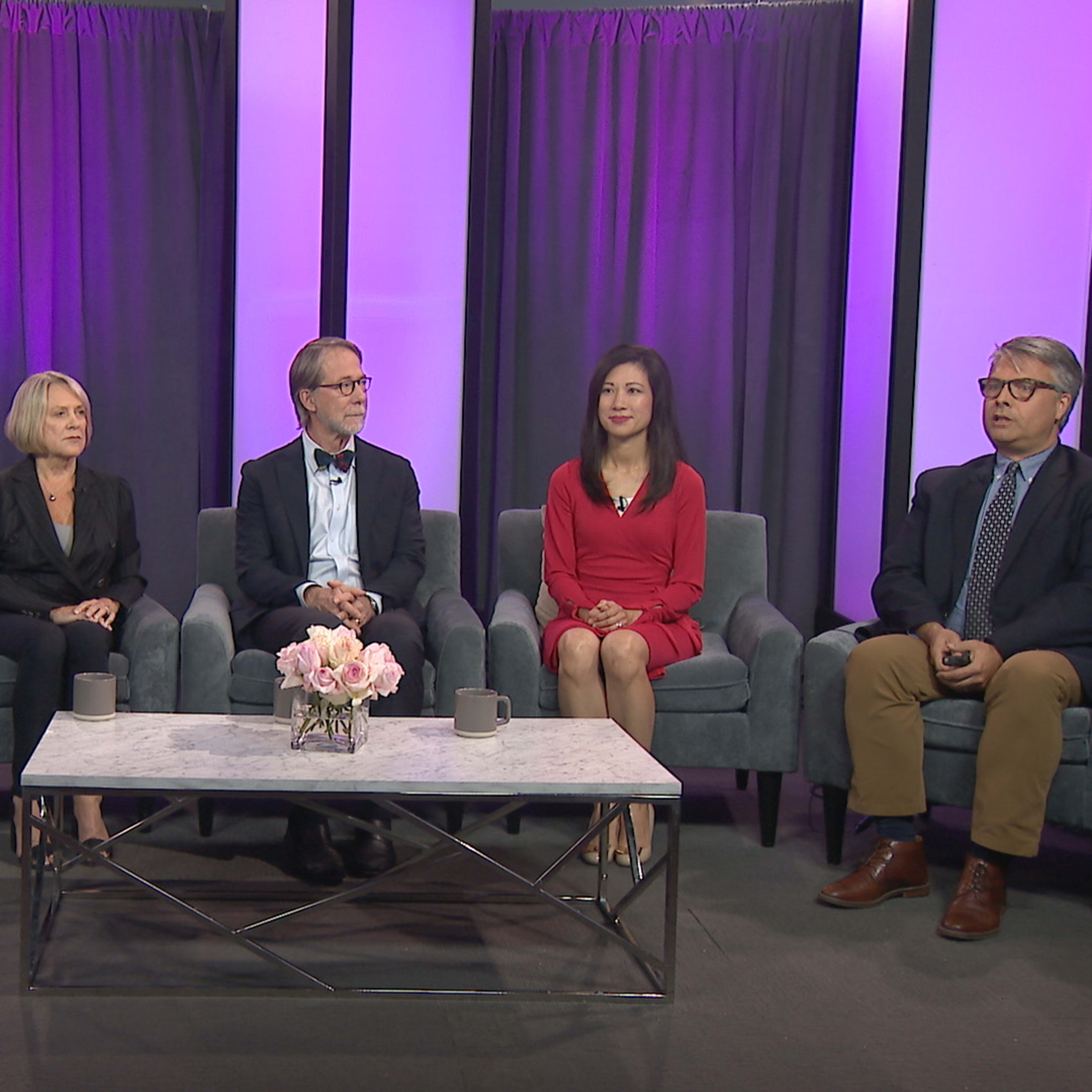 A panel of five individuals is seated on a stage with purple curtains and a table featuring a floral arrangement.