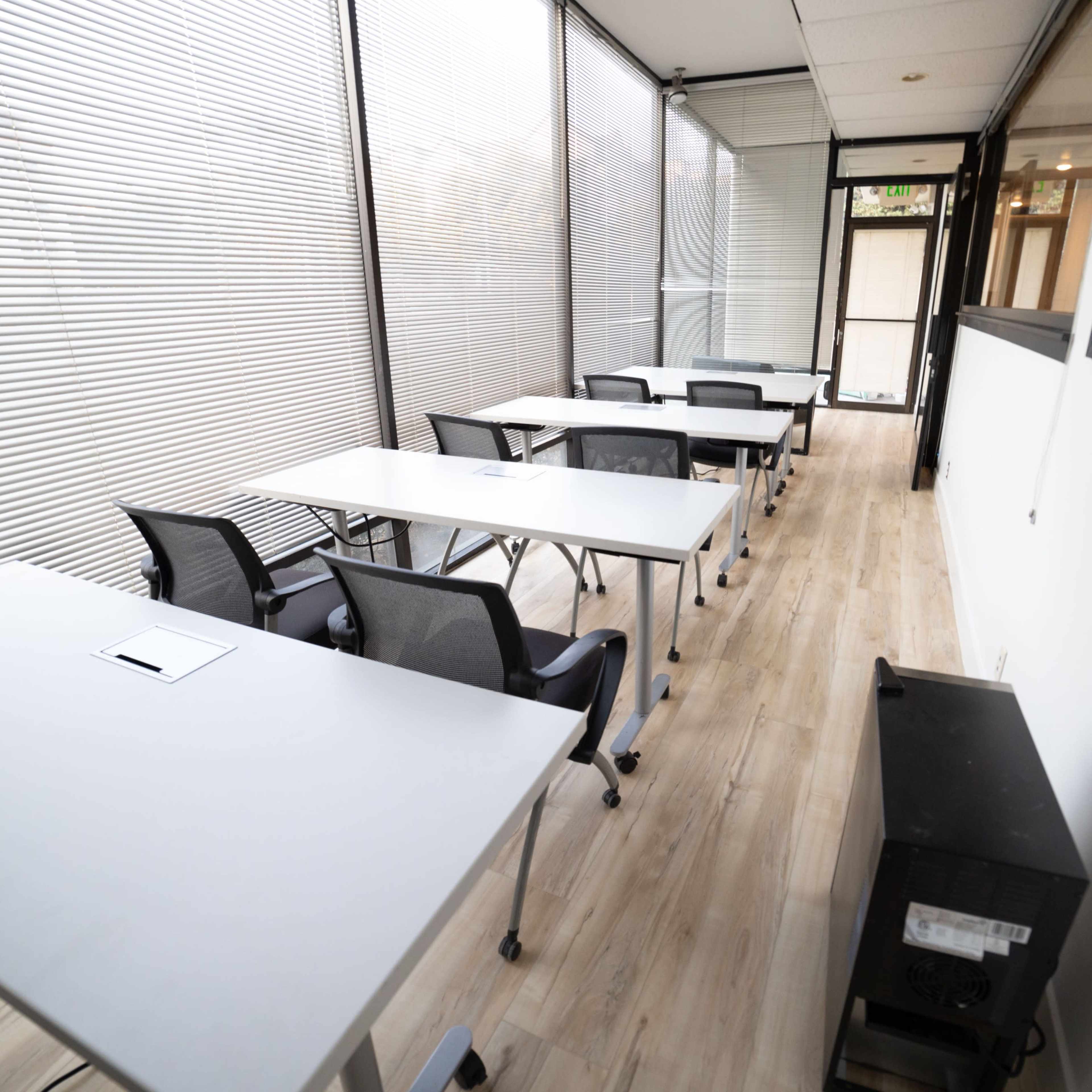 A row of tables and chairs is set up in a long, narrow classroom with large windows and light-colored wooden flooring.