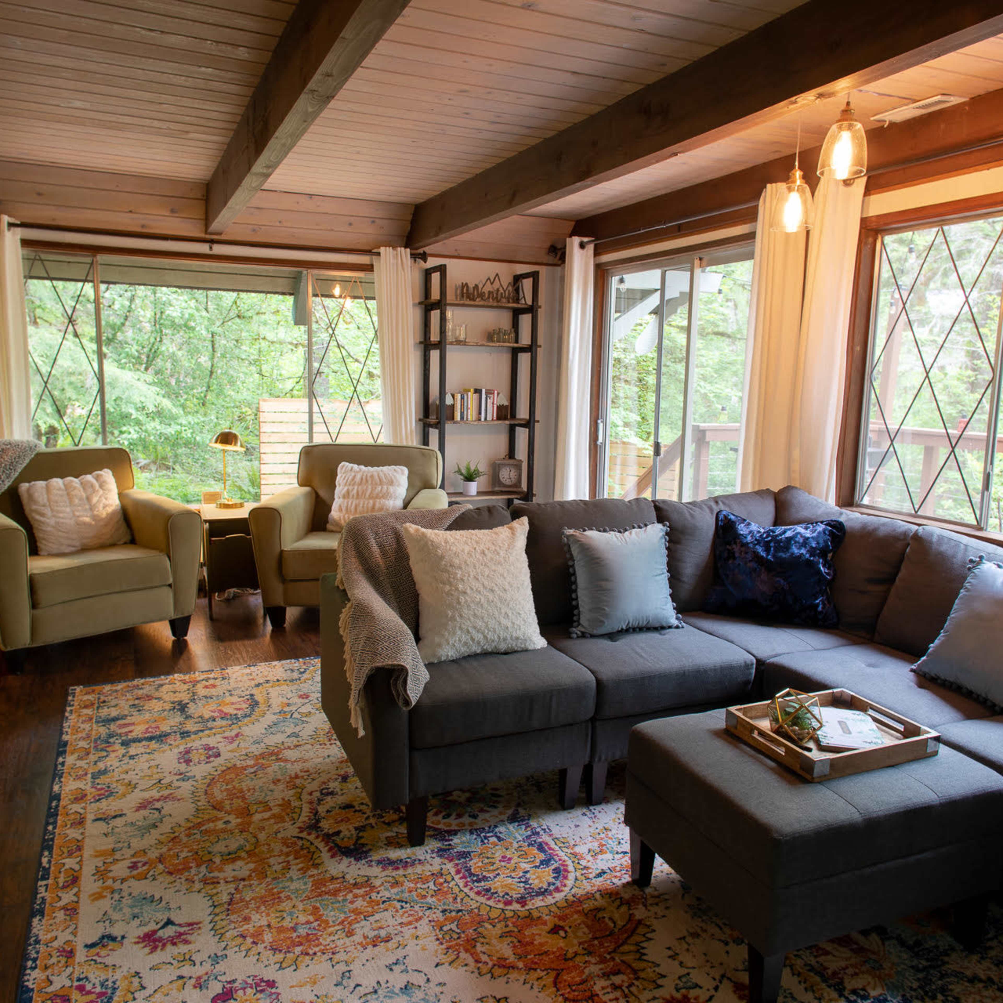 A modern living room features a gray sectional sofa, two green armchairs, and large windows overlooking a lush green landscape.