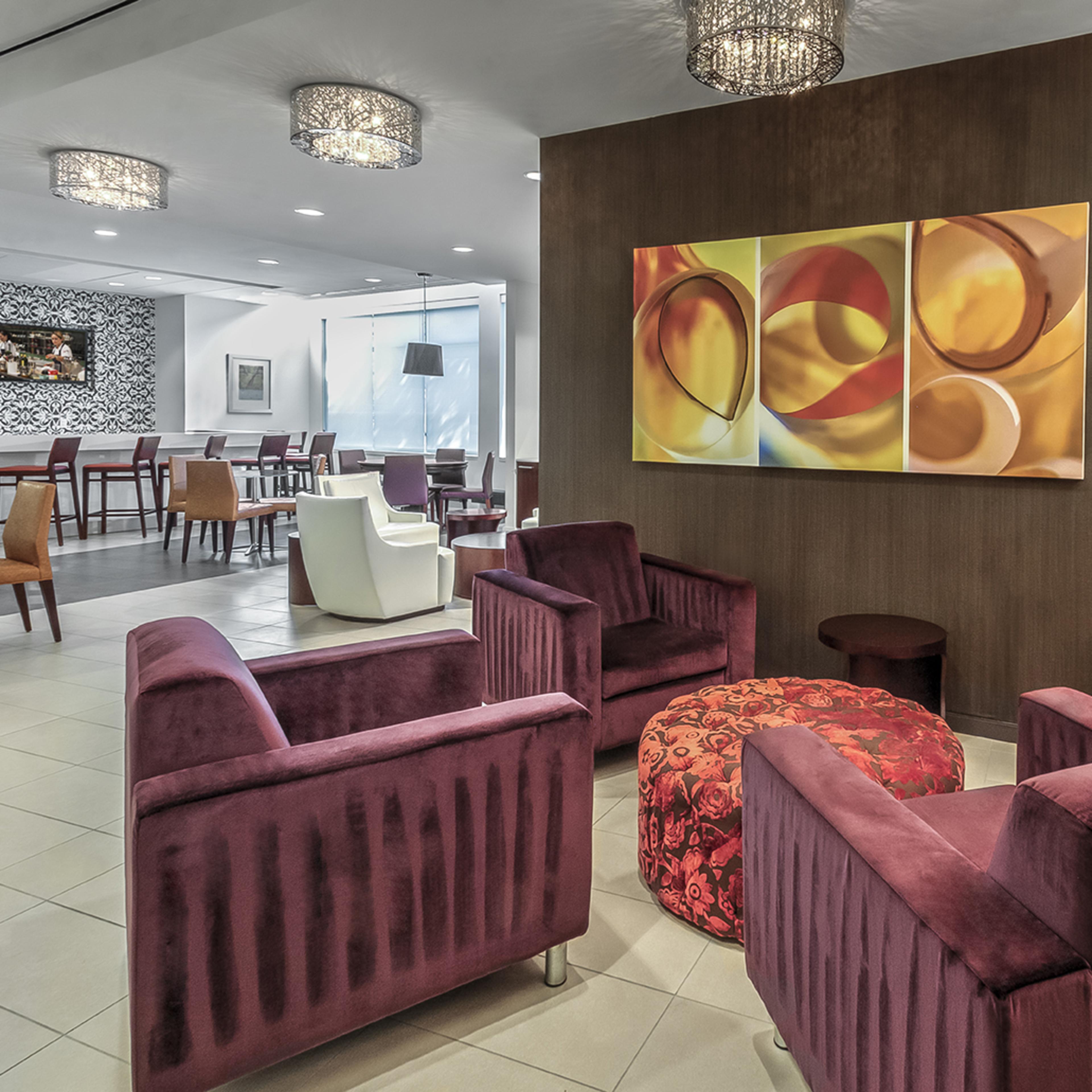 A modern lounge area with burgundy upholstered chairs, a round ottoman, and decorative lighting, adjacent to a dining space.