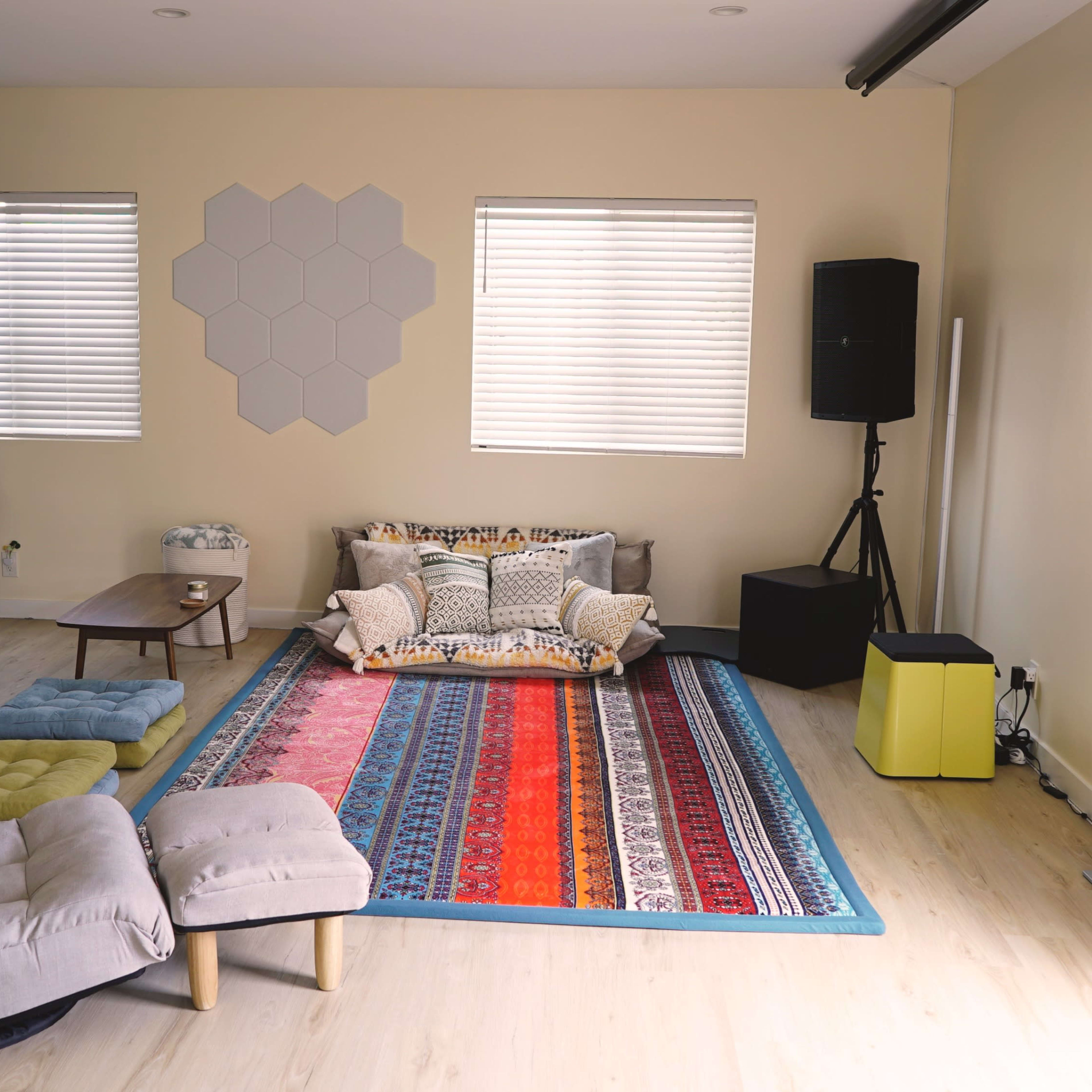 The room features a colorful area rug, a low wooden table, a sectional sofa with various cushions, a projector, and speakers, along with large windows dressed in blinds.
