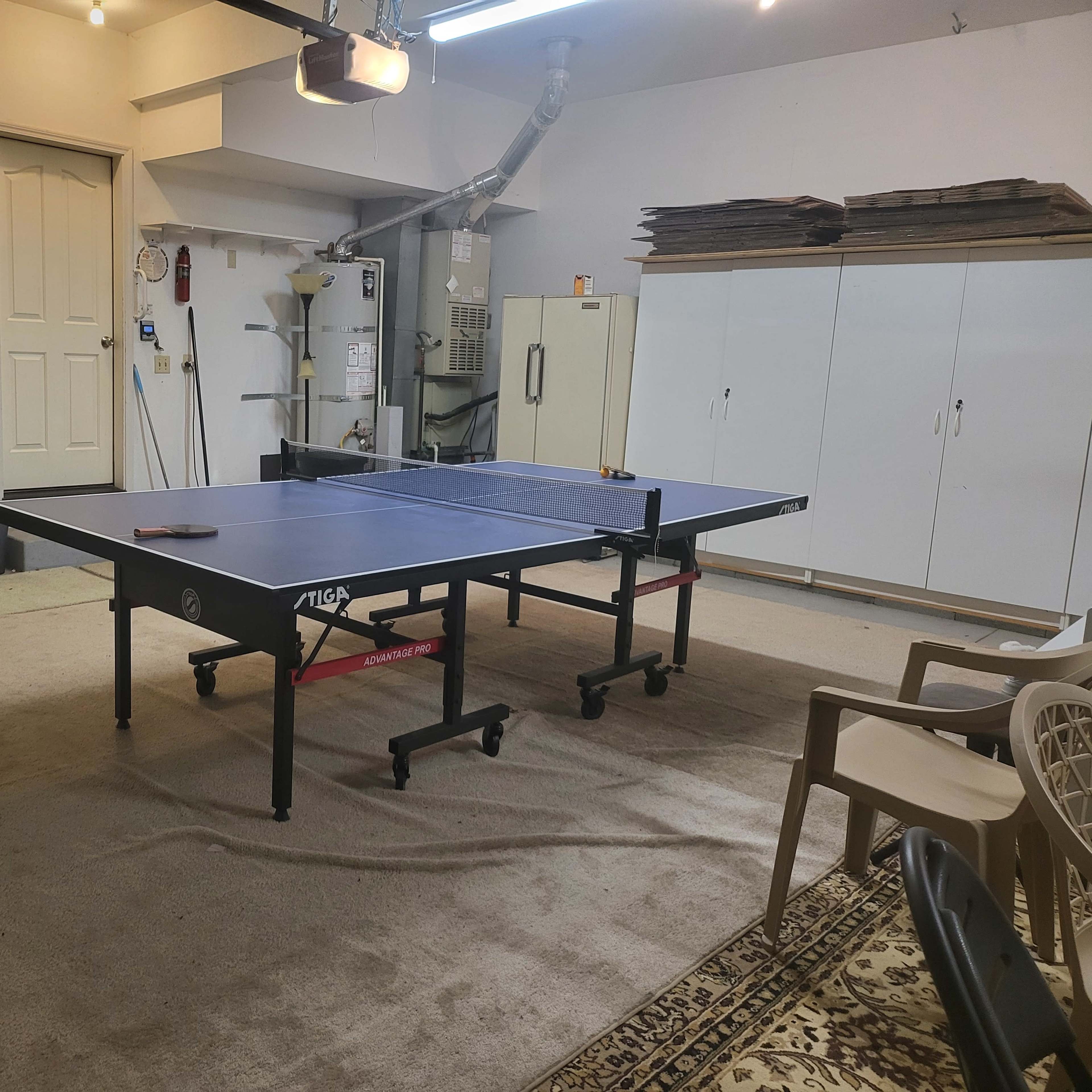 A ping pong table is set up in a garage, surrounded by chairs, cabinets, and tools.