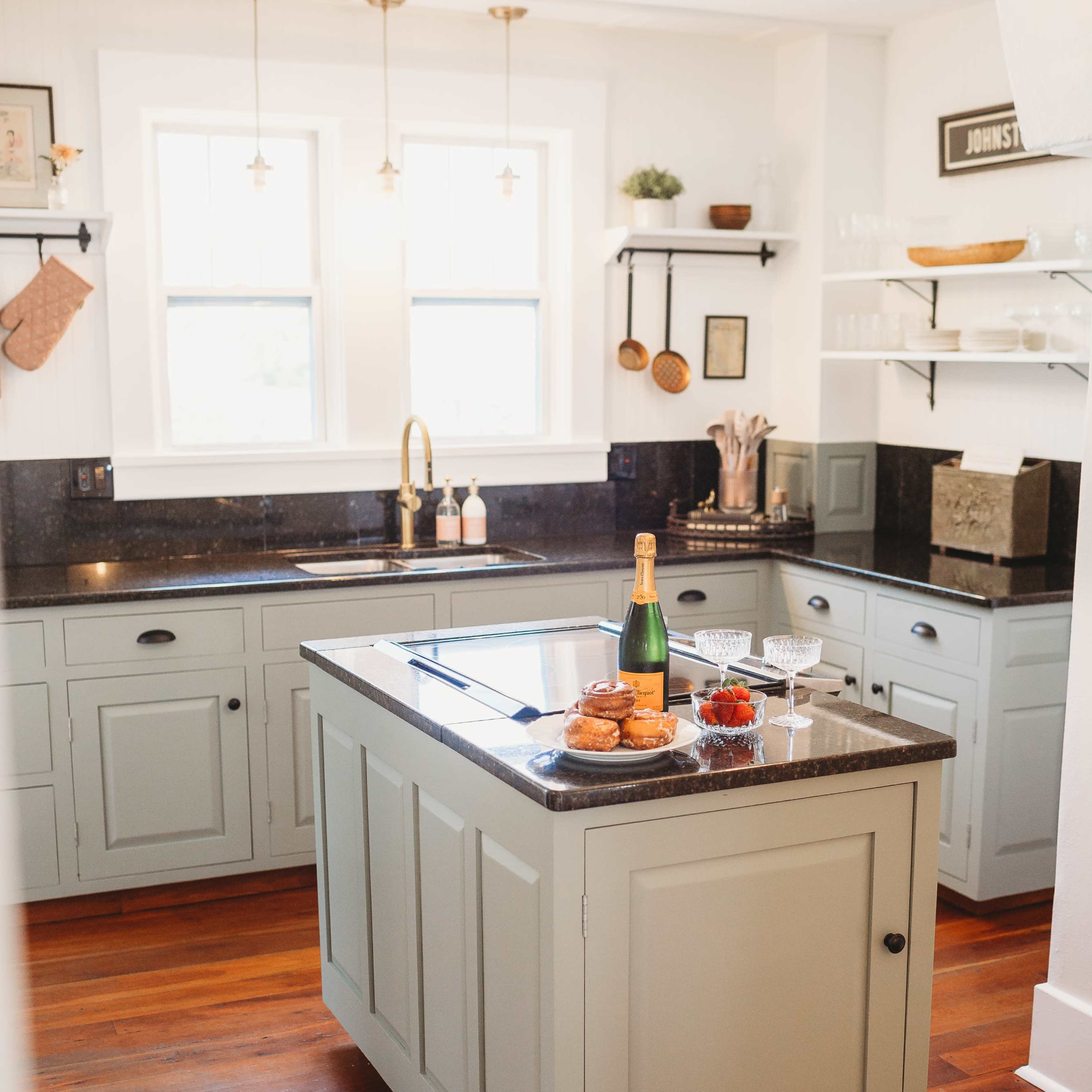 A modern kitchen features a central island with granite countertops, a bottle of champagne, a glass, and pastries, surrounded by light-colored cabinets and wooden floors.