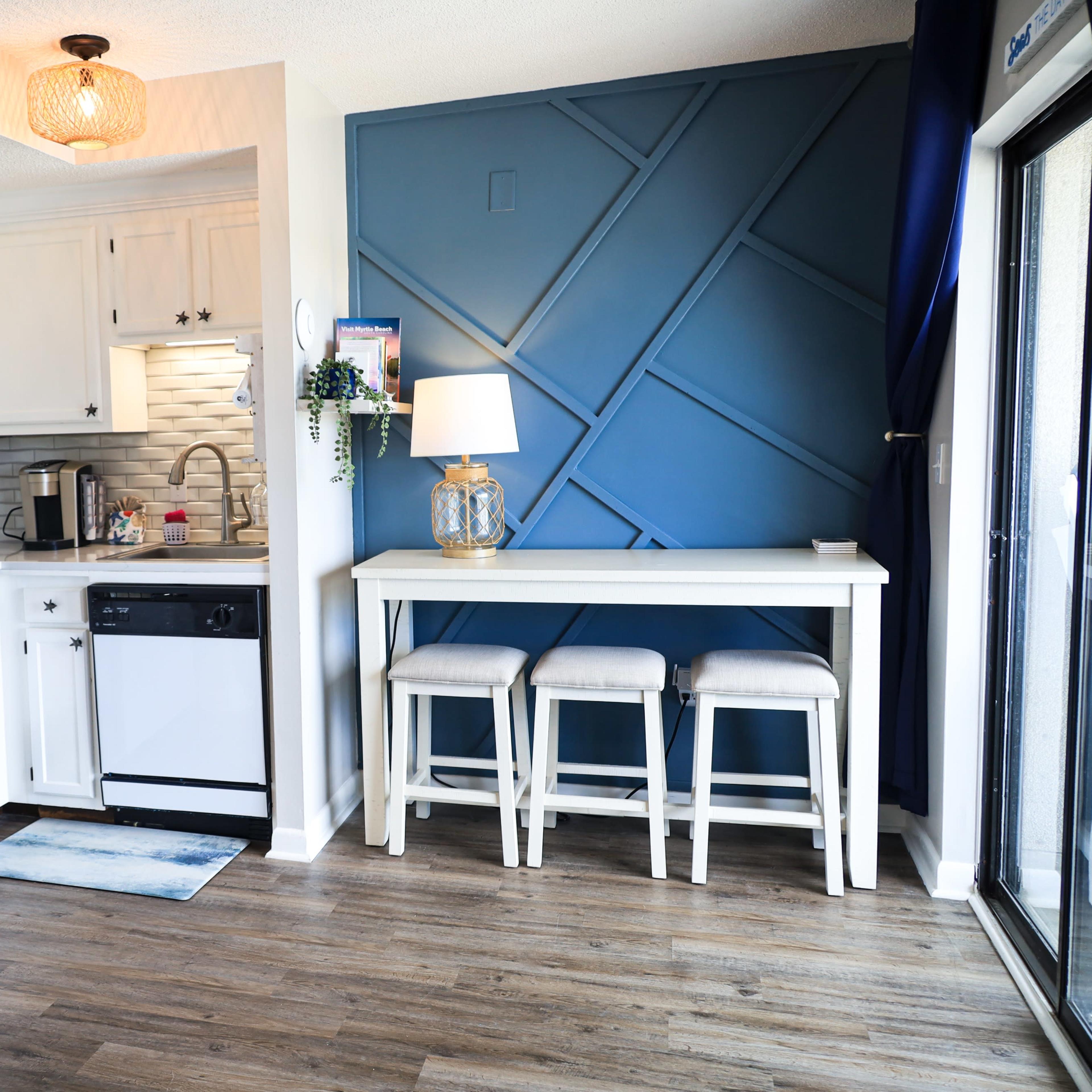 A modern kitchen and dining area feature white cabinetry, a blue accent wall, and a set of three stools at a small table.