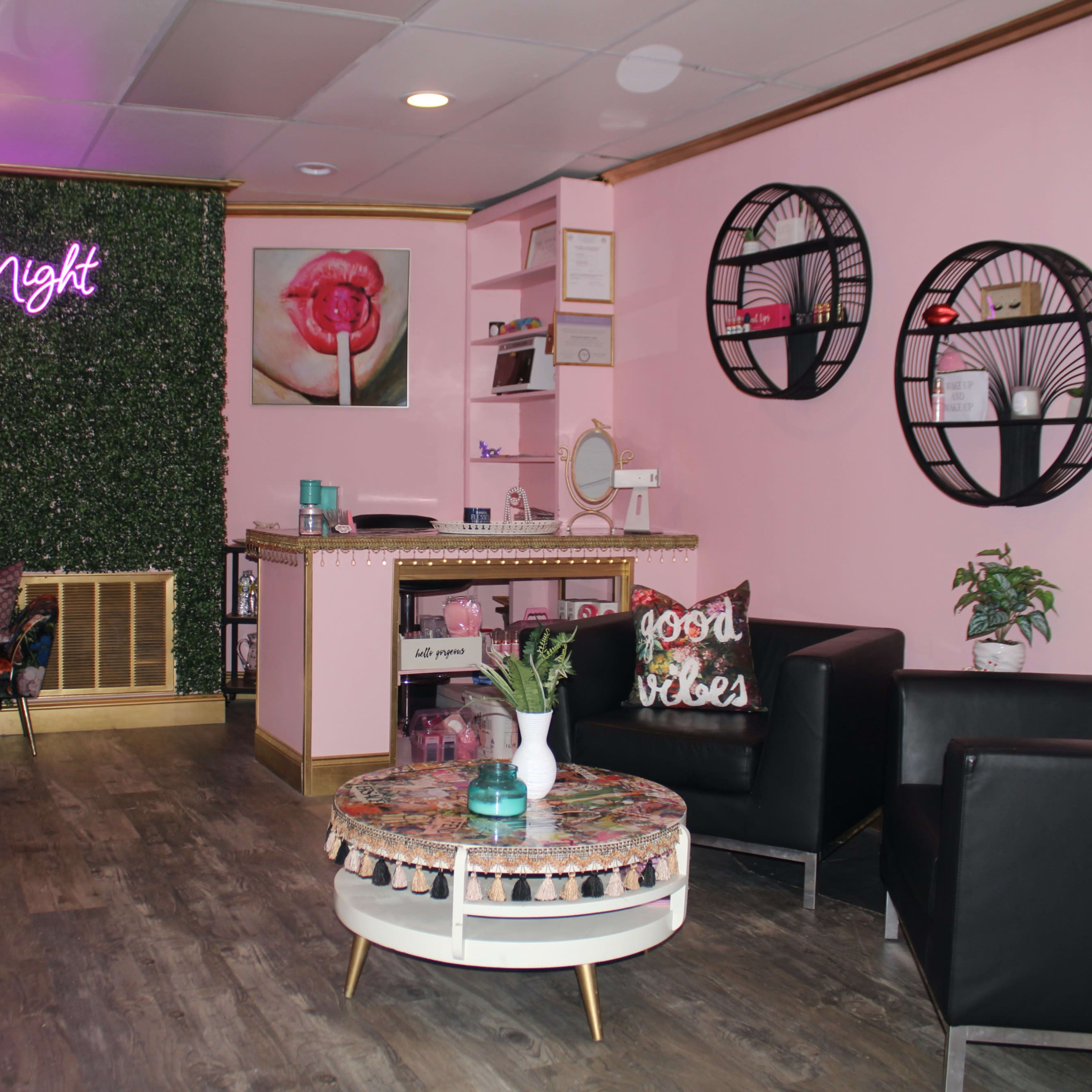 A modern beauty salon interior with pink walls, a neon sign that reads "About Lash Night," and a waiting area featuring two black chairs, a decorative coffee table, and wall decor.