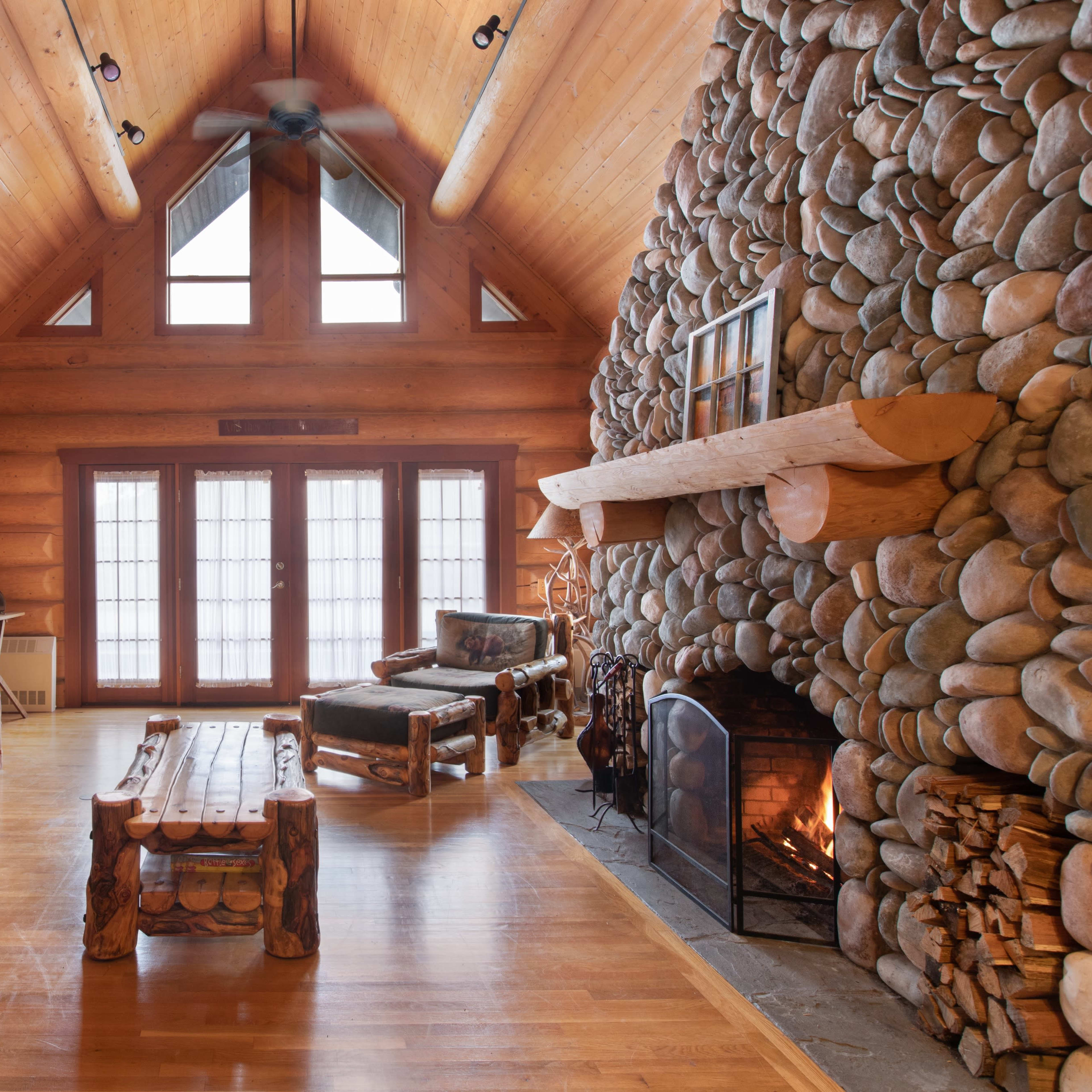 The interior of a log cabin features a stone fireplace, wooden furniture, and large windows that provide natural light.