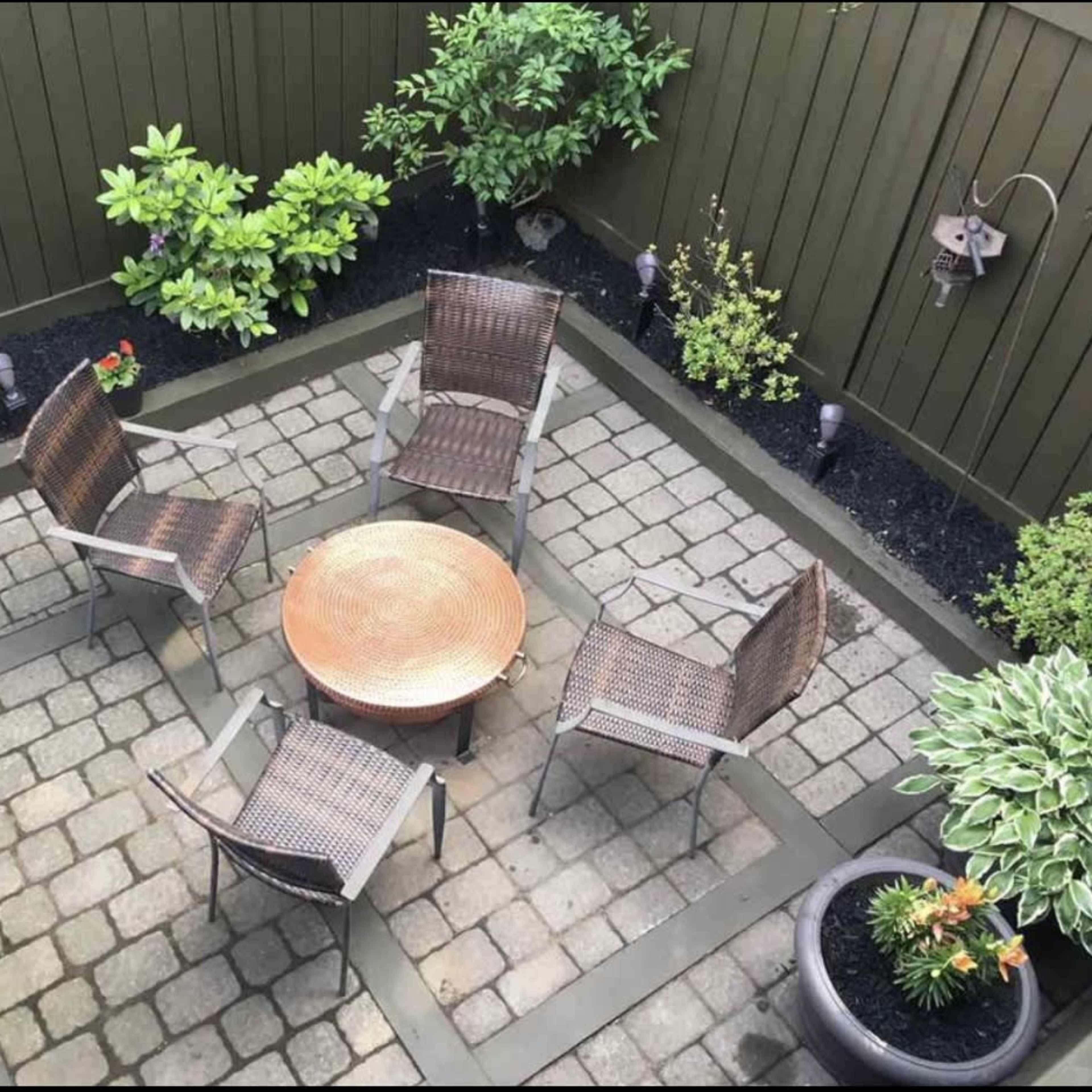 A small outdoor patio with four wicker chairs arranged around a round metal table, surrounded by greenery and potted plants.