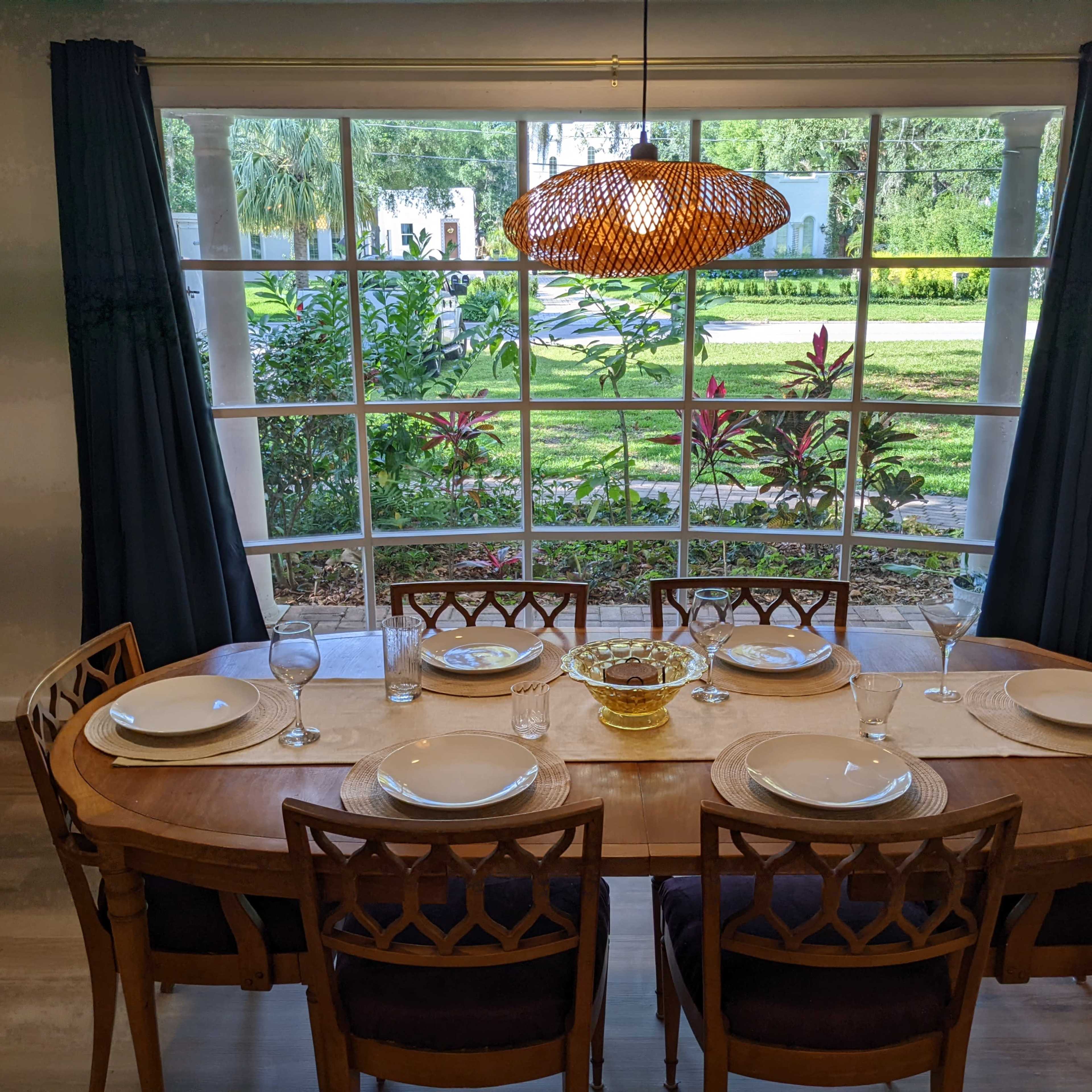 A round dining table is set with plates and glasses, positioned near a large window overlooking a garden.