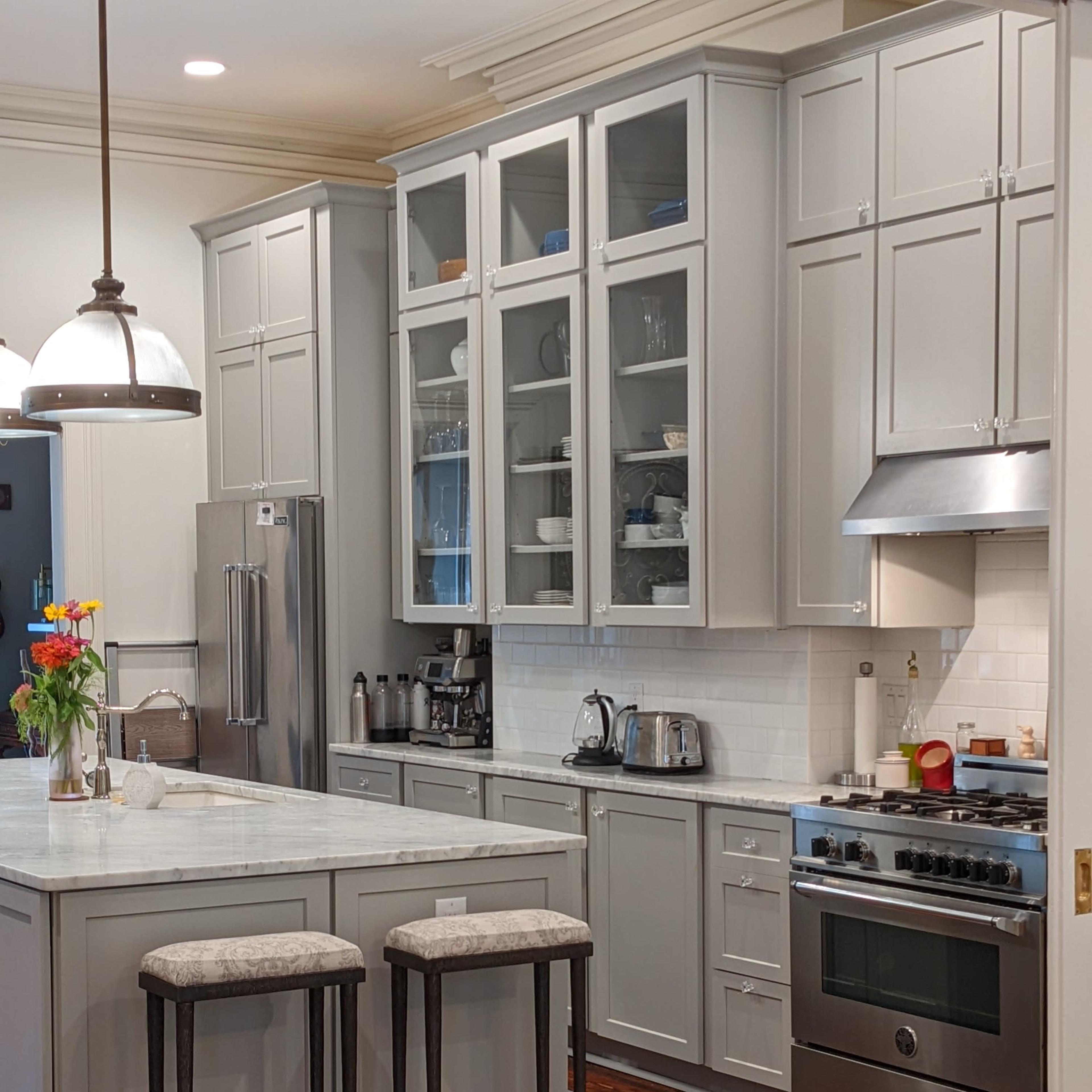 A modern kitchen features gray cabinetry, a marble countertop island with two stools, and stainless steel appliances.