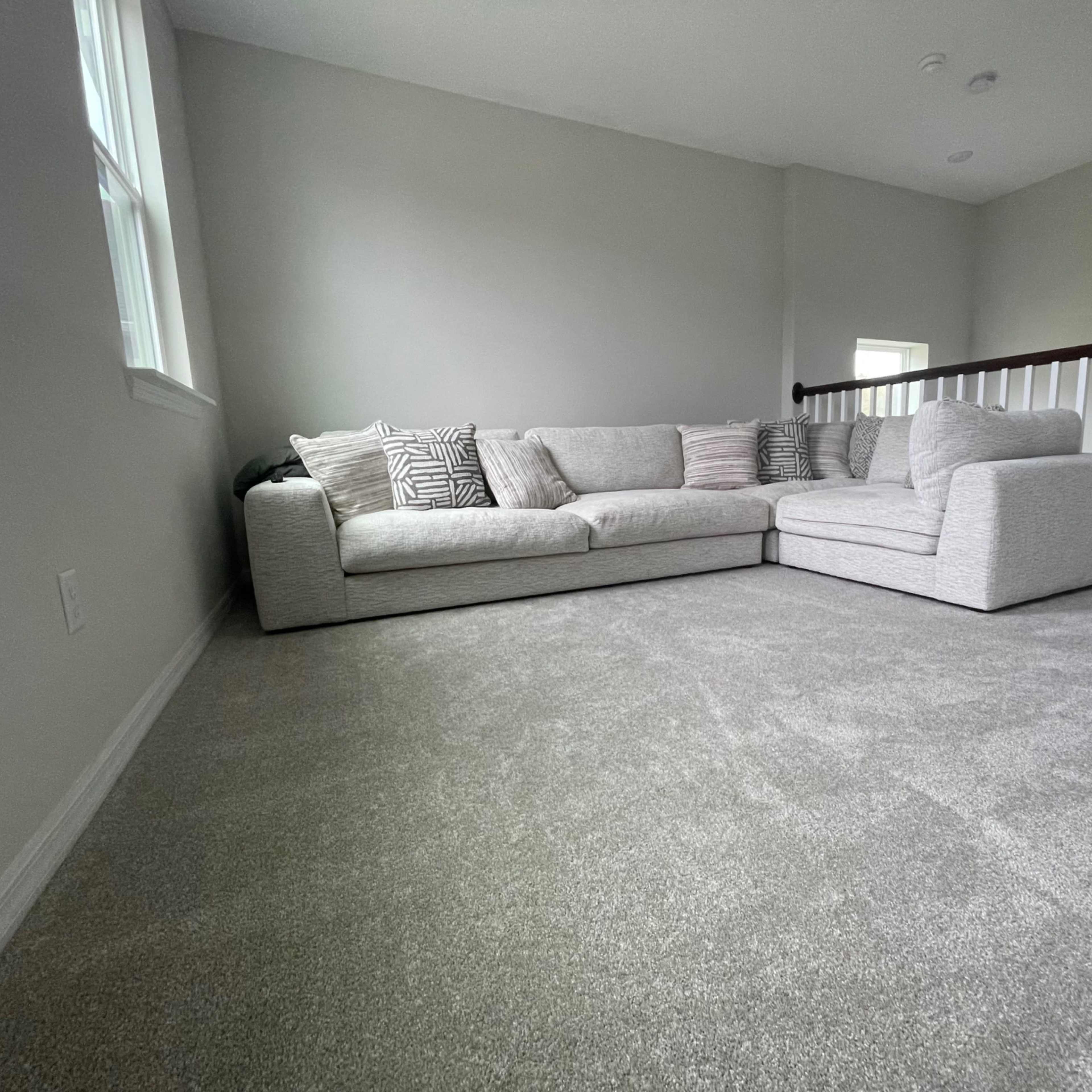 A large sectional sofa with decorative pillows is positioned in a carpeted room with light gray walls and a railing visible in the background.