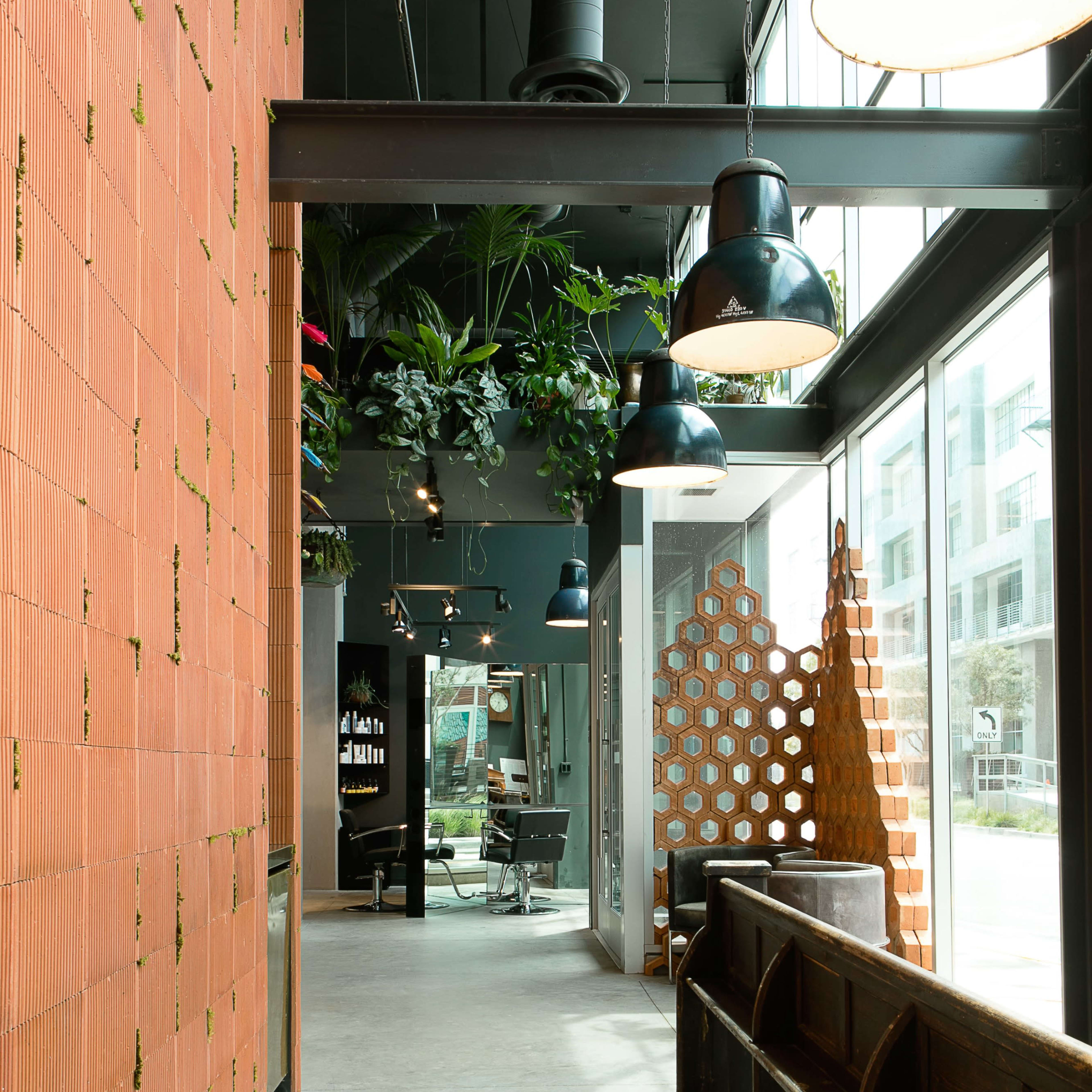 A modern interior hallway featuring a textured orange brick wall, hanging lights, and lush greenery, with a seating area and decorative wooden partition at the far end.