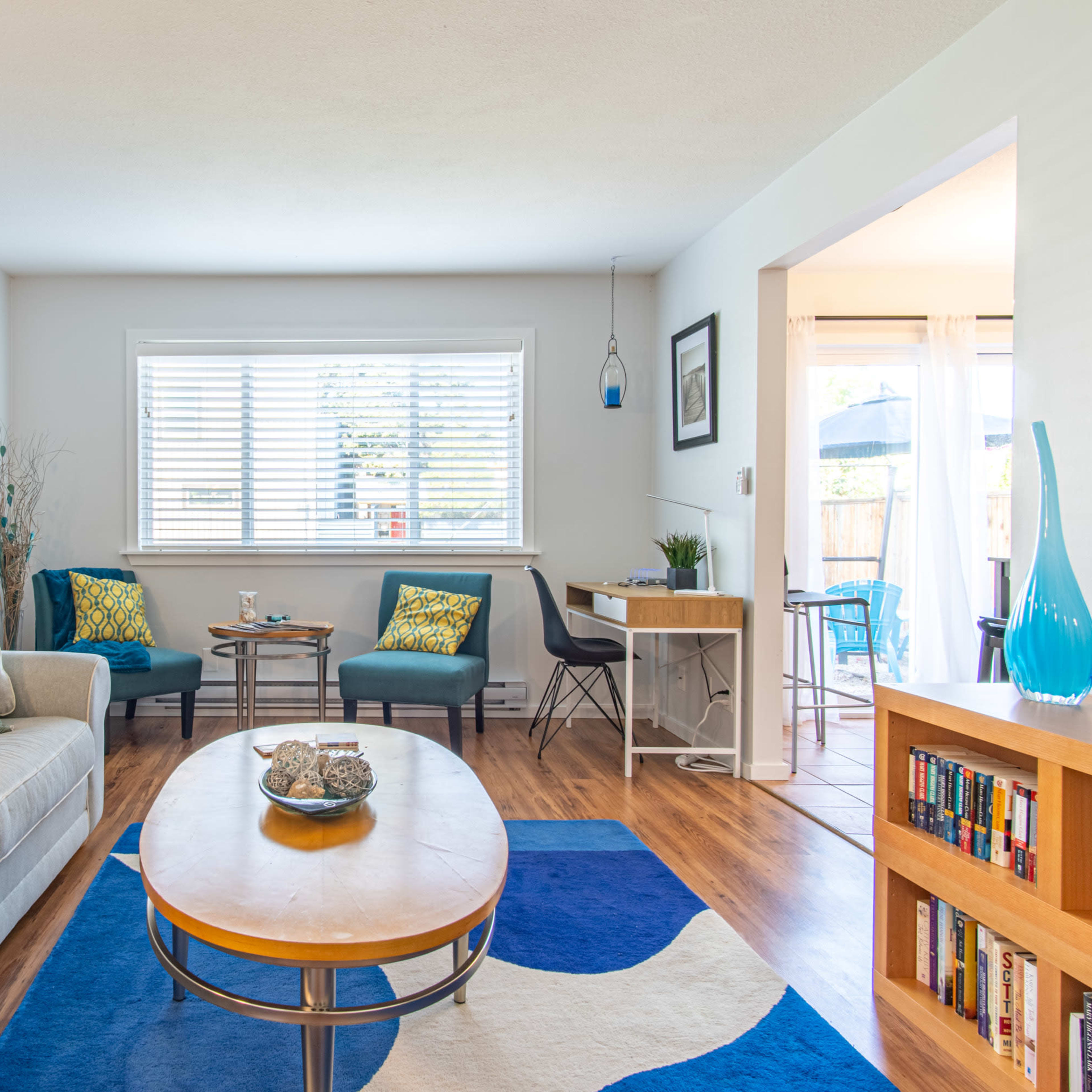 A bright living room with a sofa, a wooden coffee table, and two chairs beside a window, complemented by a small dining area in the background.