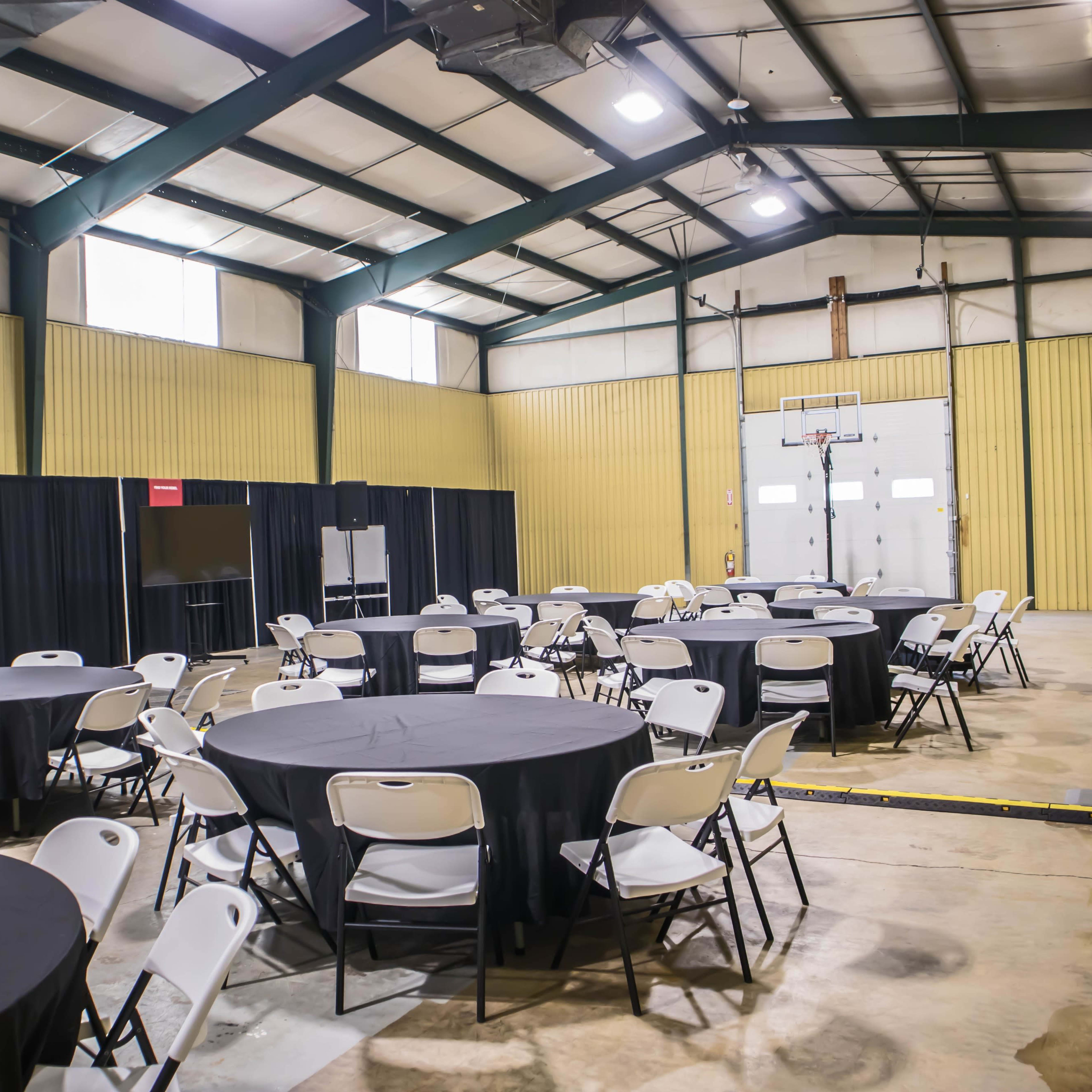 A spacious indoor hall features circular tables surrounded by chairs, with black drapes and a basketball hoop at the far end.
