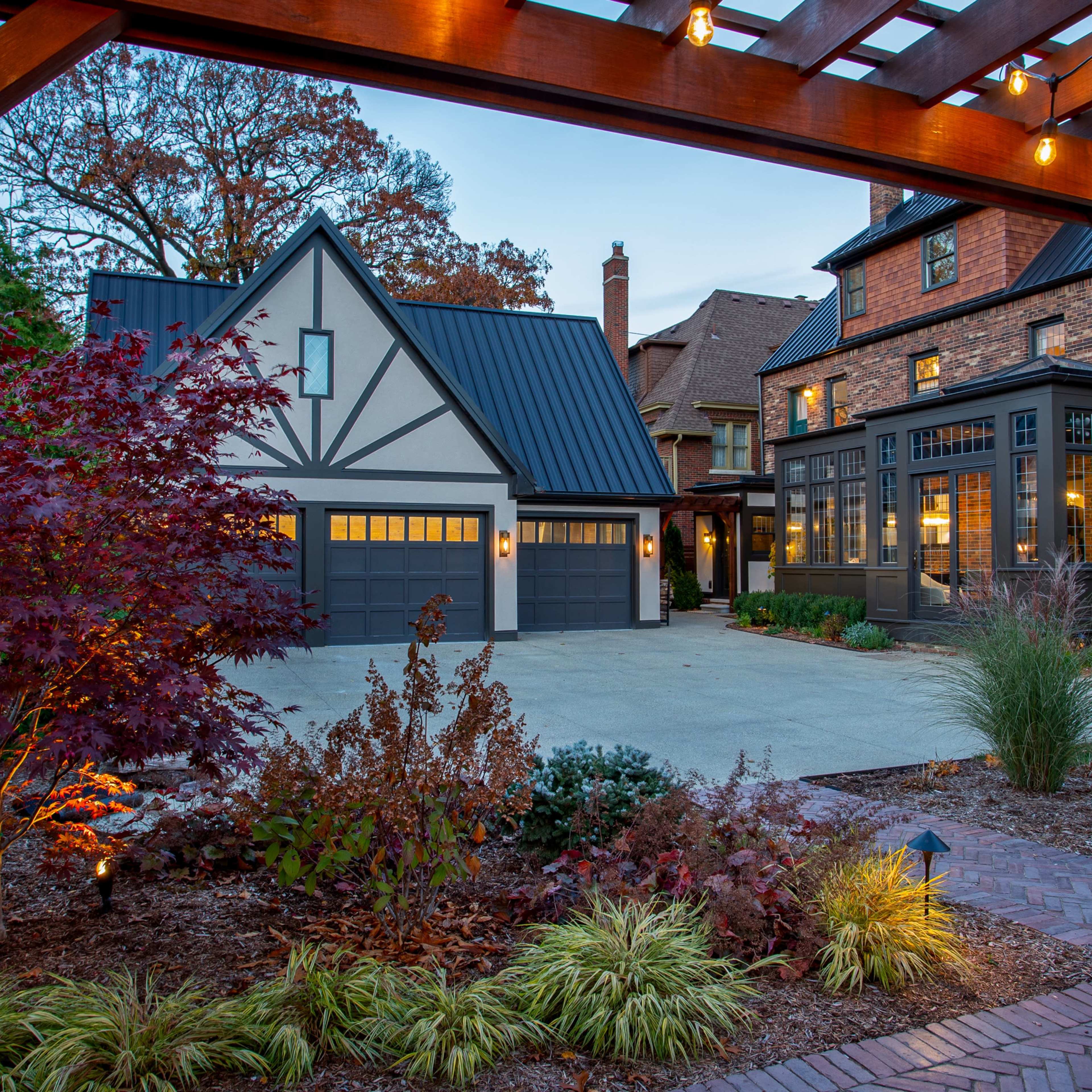A landscaped residential area featuring two distinct houses, a garage with a modern roof, and outdoor lighting under a wooden pergola.