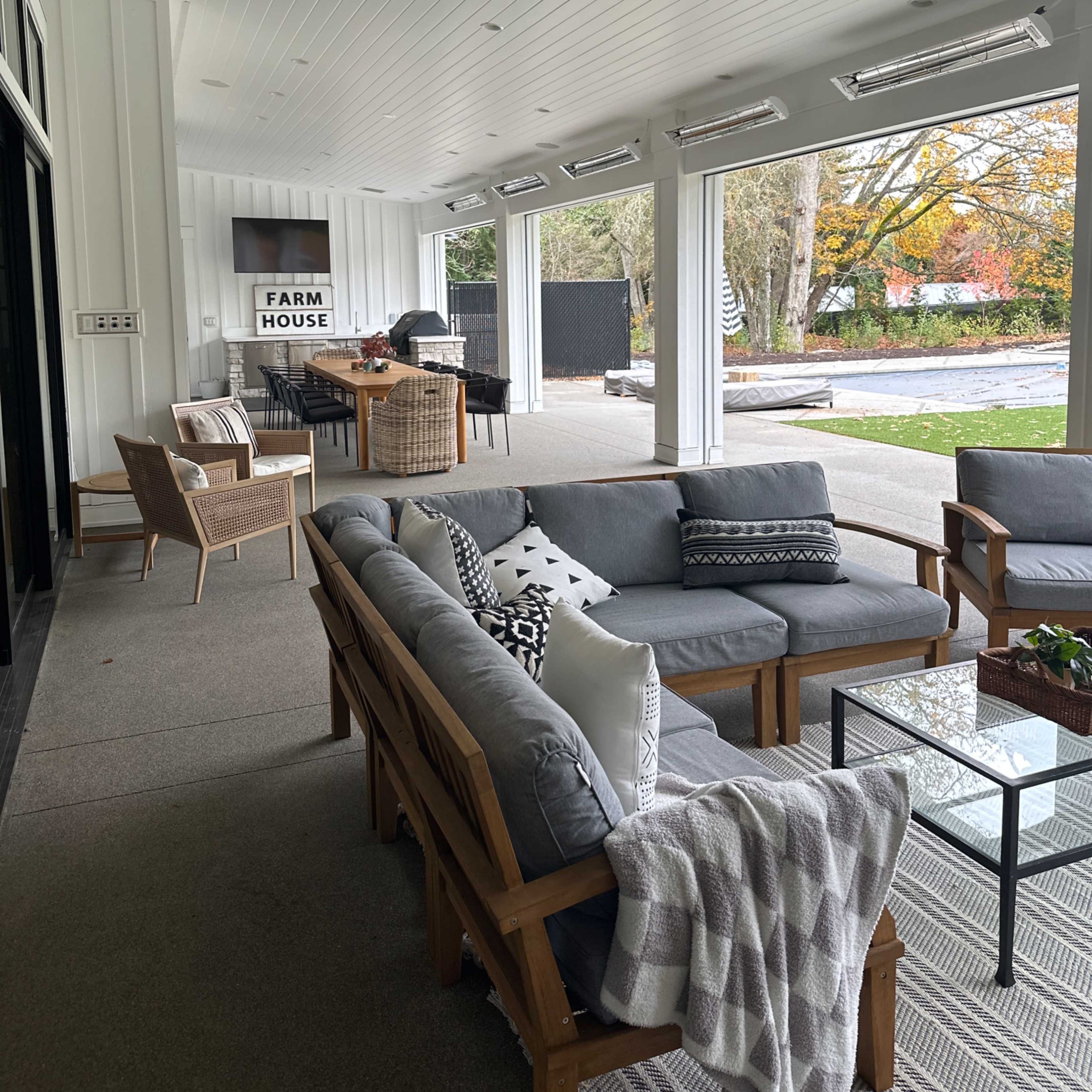 A modern covered patio with a seating area, a dining space, and a barbecue setup, featuring white walls and large windows.