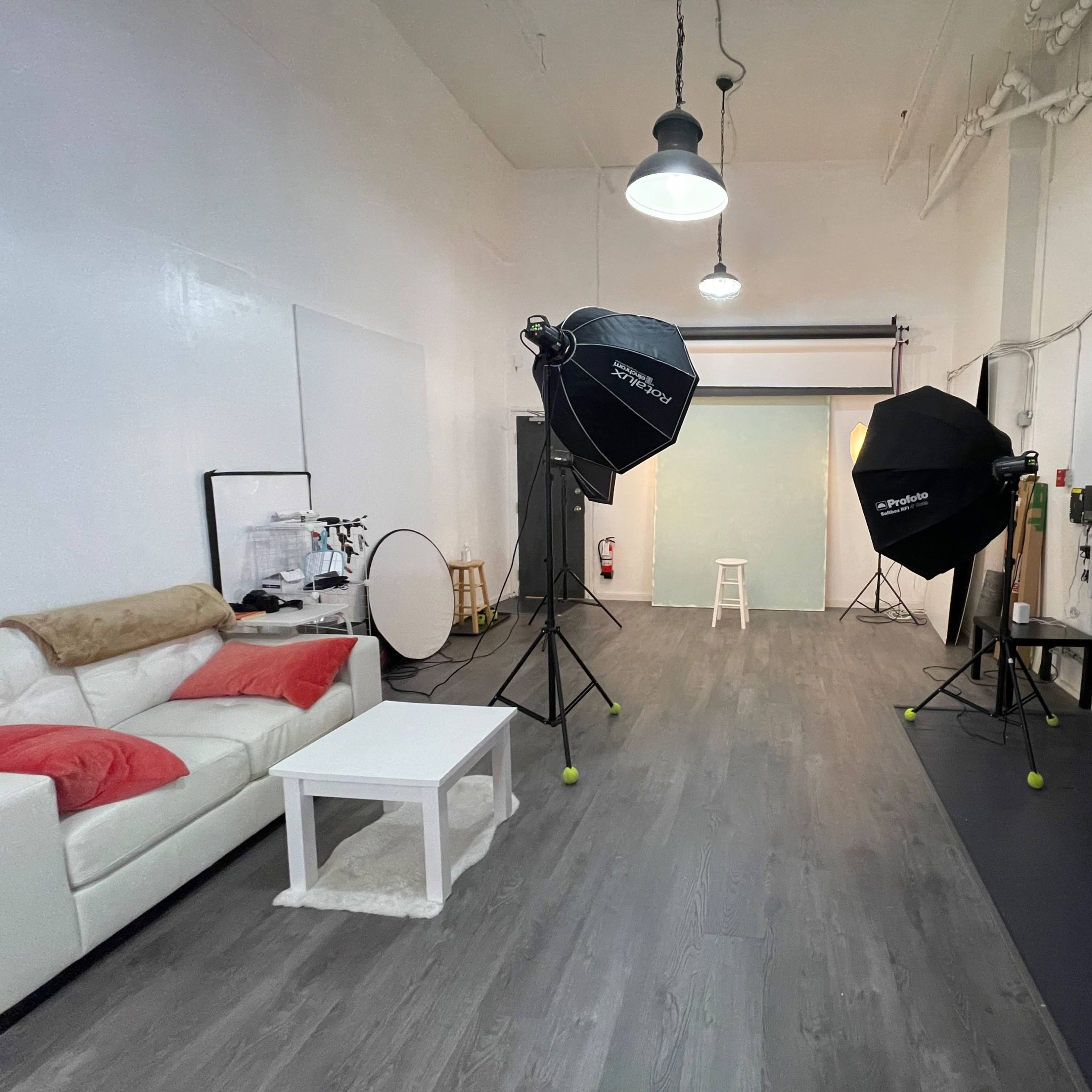 A photography studio features a white wall, wooden flooring, a white couch with red pillows, studio lights, and a backdrop setup.
