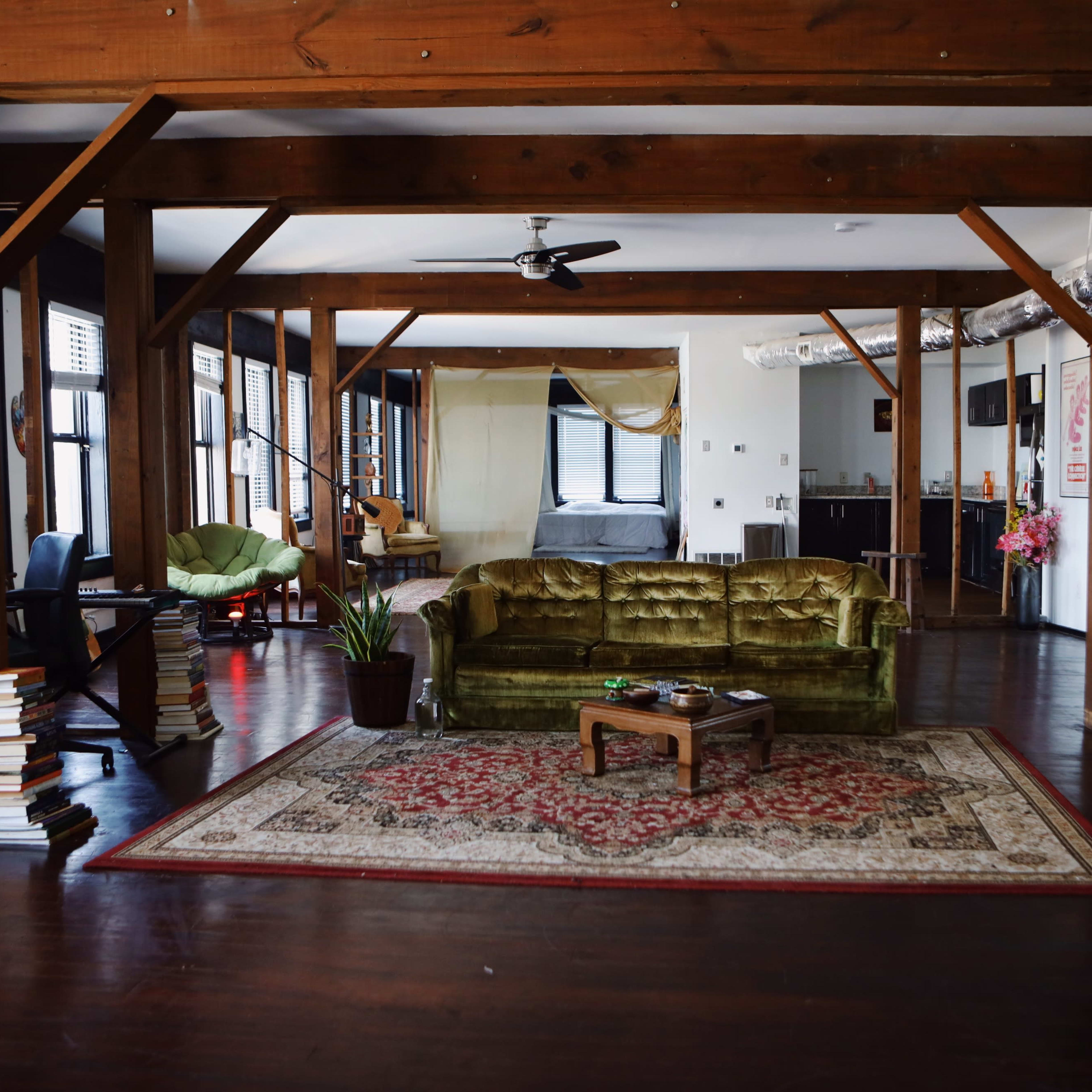 A spacious loft-style living room featuring wooden beams, a green velvet couch, a coffee table, and a bookshelf stacked with books.
