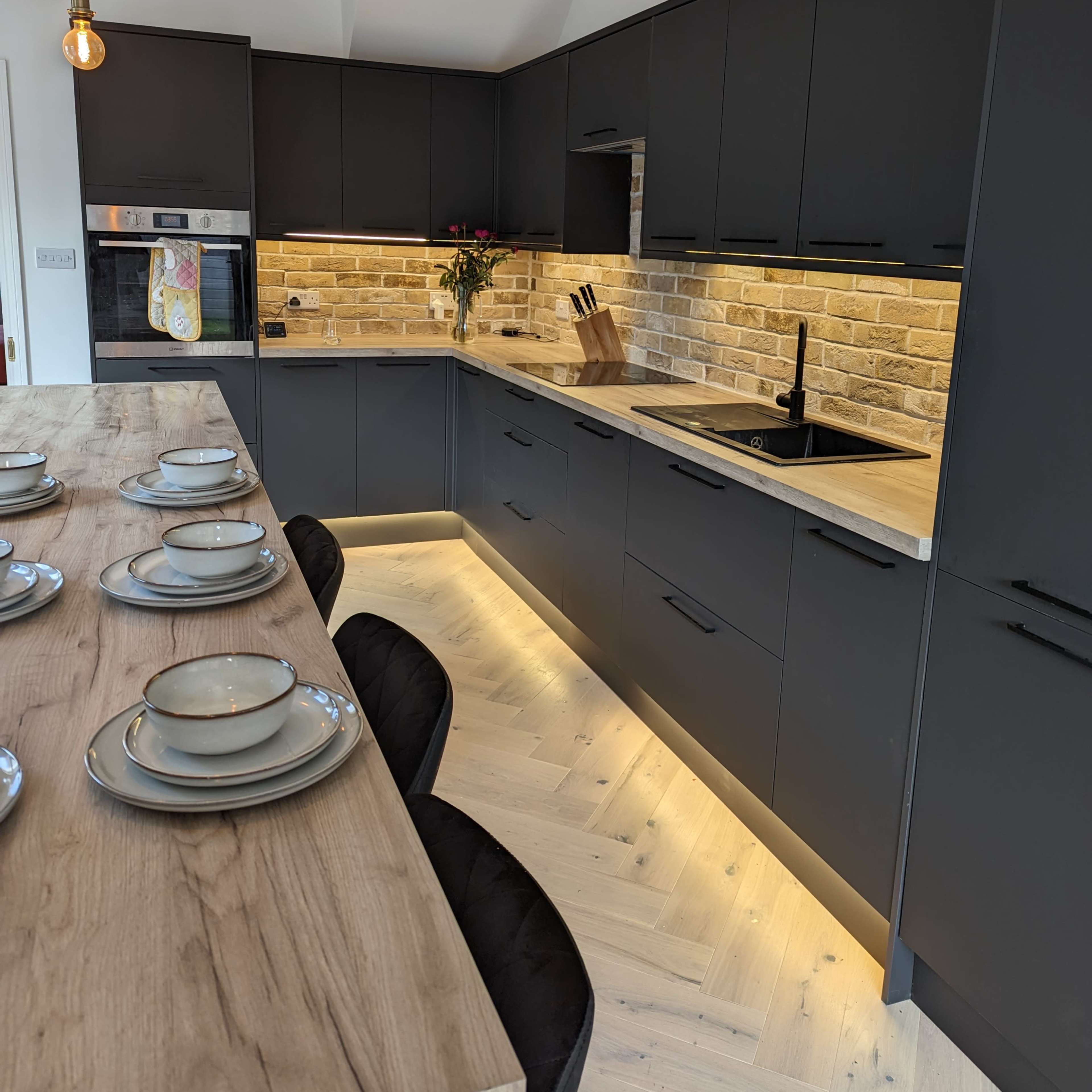 A modern kitchen with dark cabinetry, a wood countertop, and under-cabinet lighting illuminating the stone backsplash.