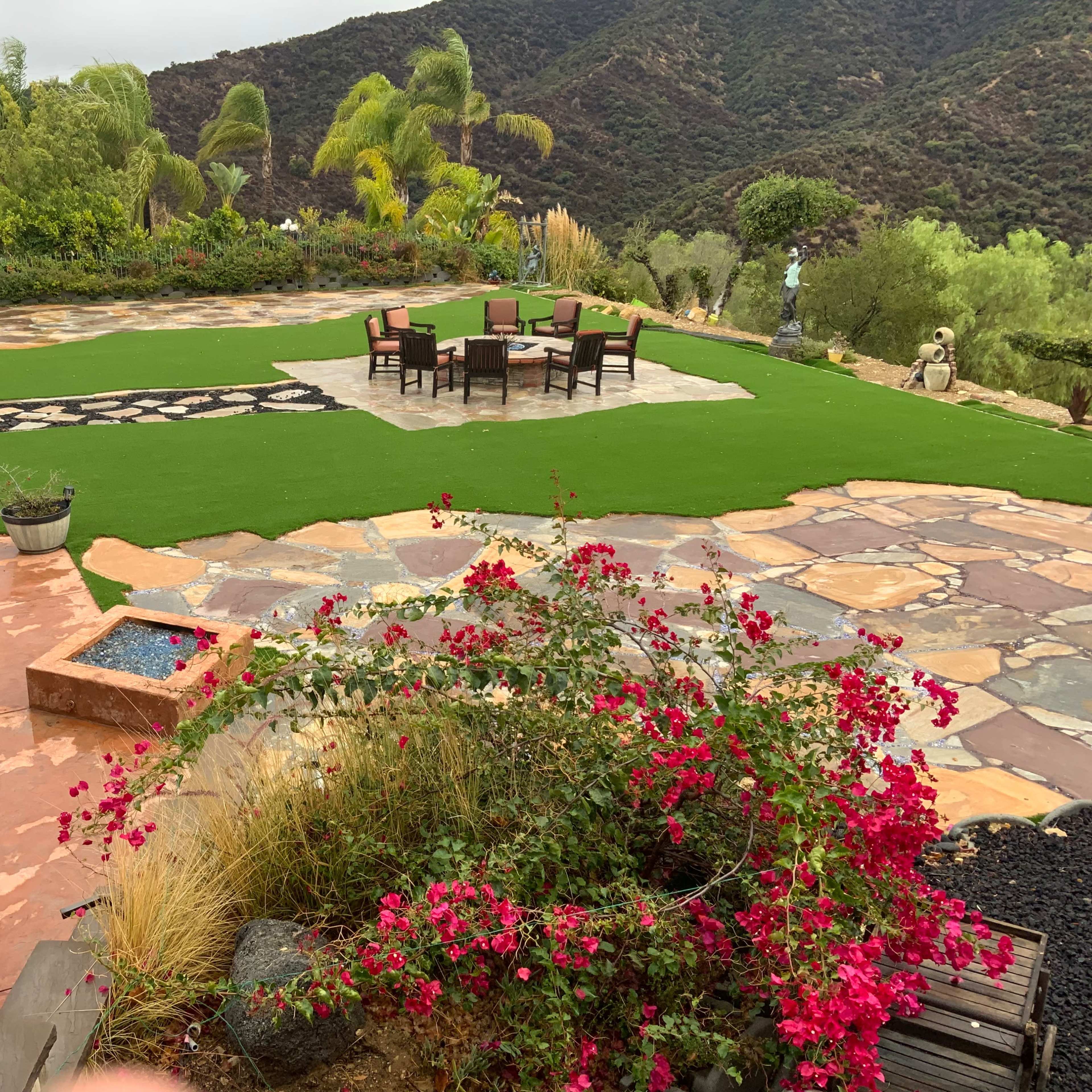 A landscaped outdoor area featuring a stone patio, a green lawn, a dining set, and blooming bougainvillea in the foreground, with hills in the background.
