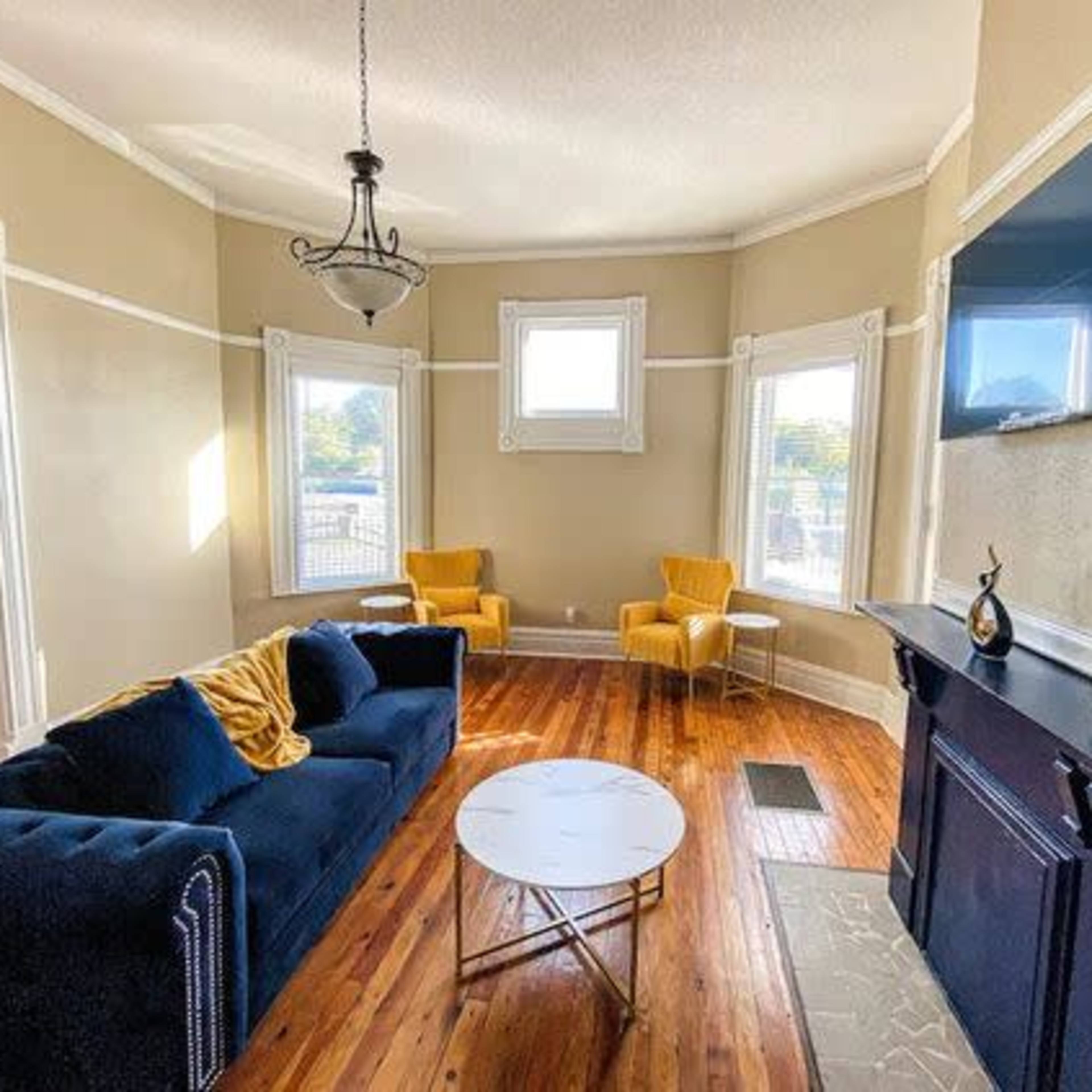 A living room featuring a blue sofa, a round coffee table, two yellow chairs, and a wall-mounted television.
