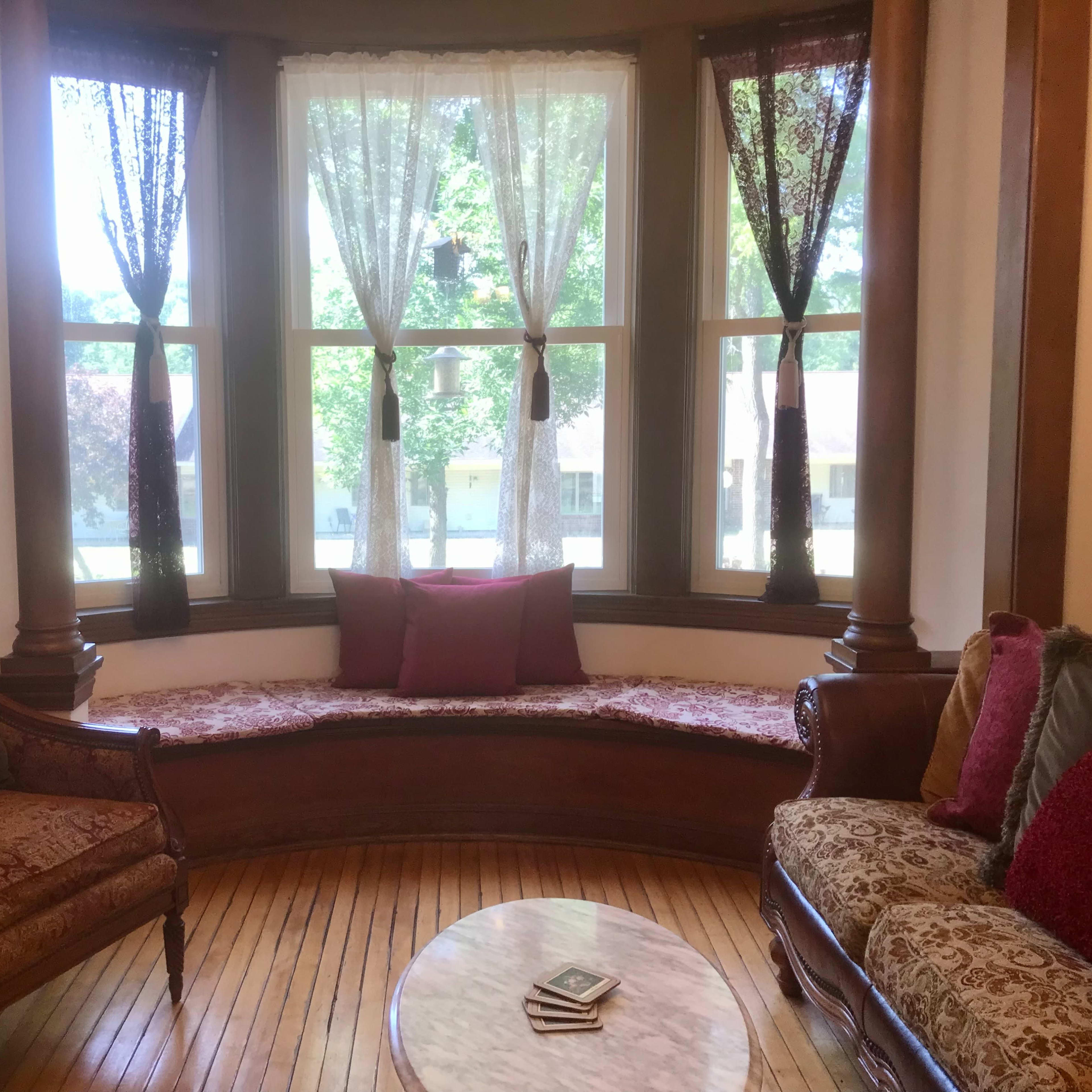 A brightly lit bay window area with a cushioned bench, surrounded by two upholstered chairs and a small round table with a stack of books.