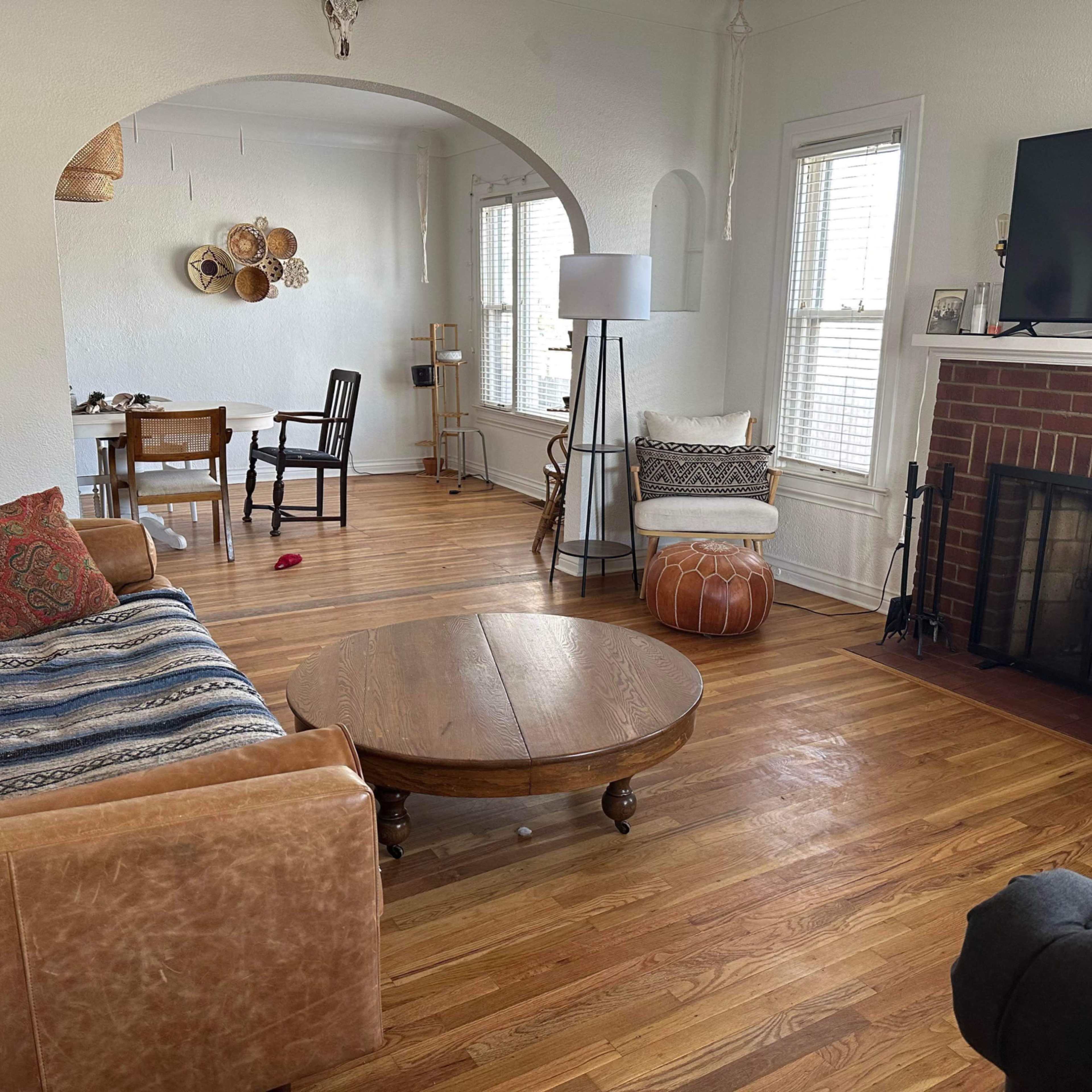 A living room with a brown leather sofa, a round wooden coffee table, a television mounted on the wall, and a fireplace surrounded by wooden flooring and various furnishings.