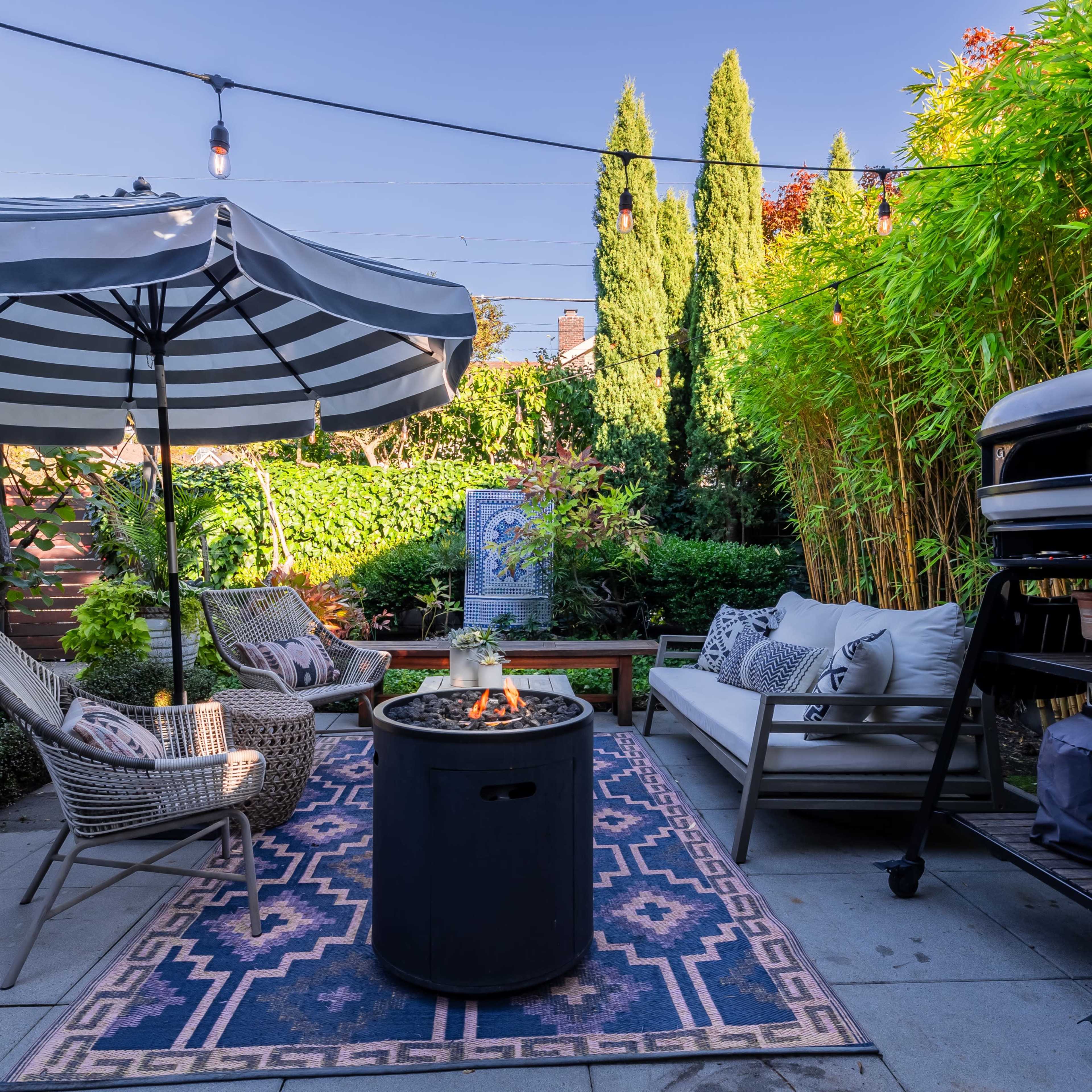 A backyard seating area features a striped umbrella over chairs, a fire pit on a patterned rug, and a pizza oven nearby, surrounded by greenery.