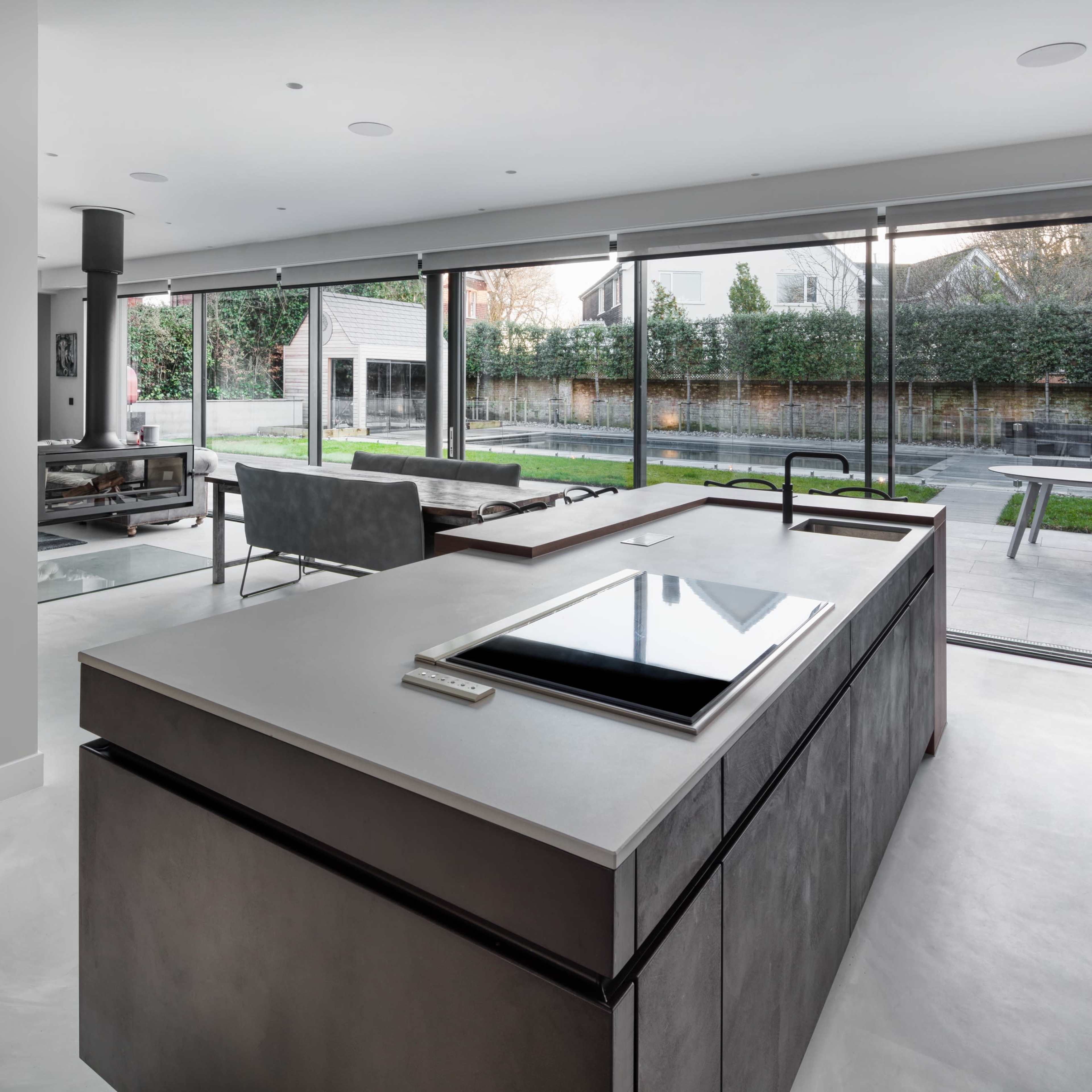 A modern kitchen with a large island, featuring a cooktop, overlooking a spacious outdoor area through expansive glass windows.