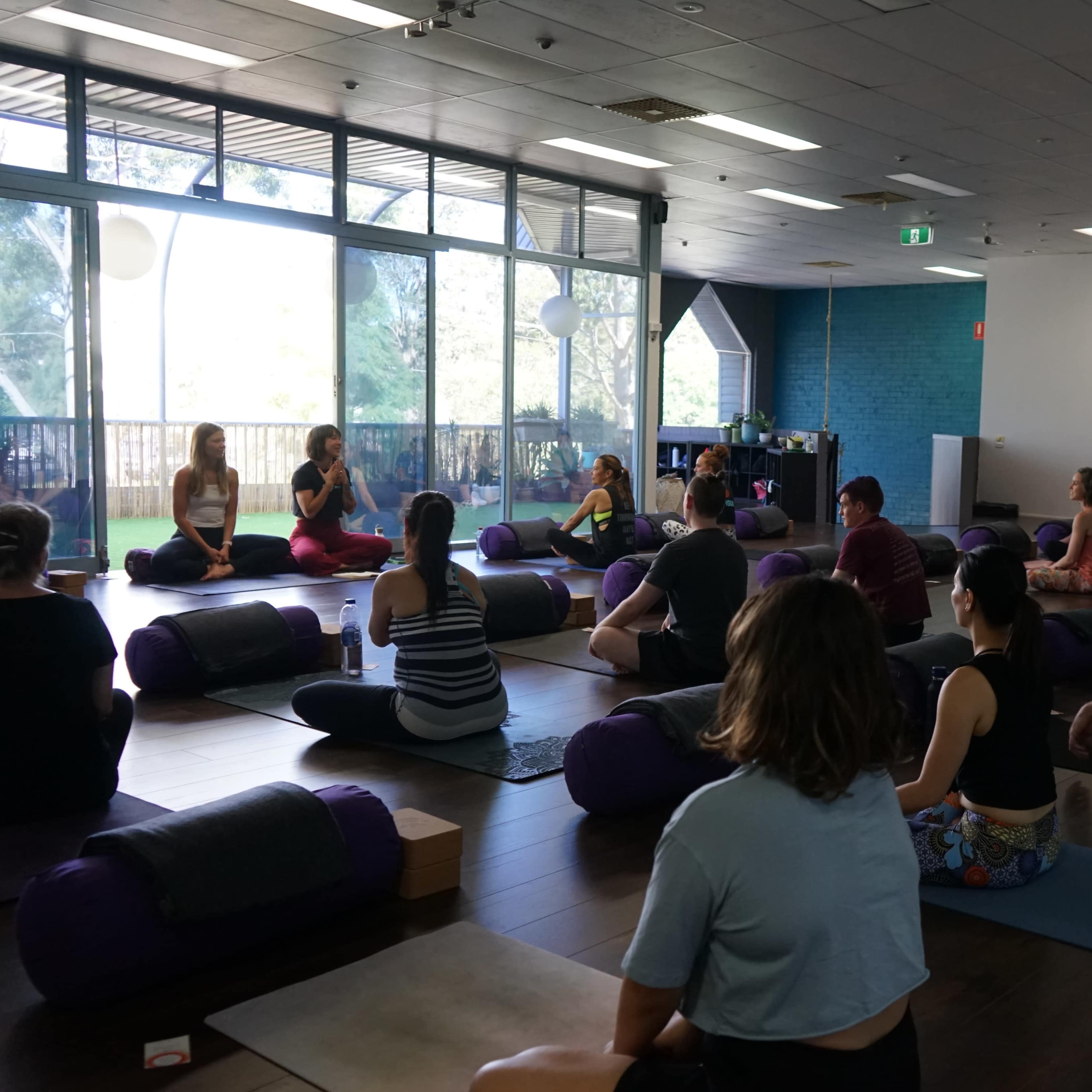 A group of people participate in a yoga class, seated on mats and cushions in a well-lit studio.