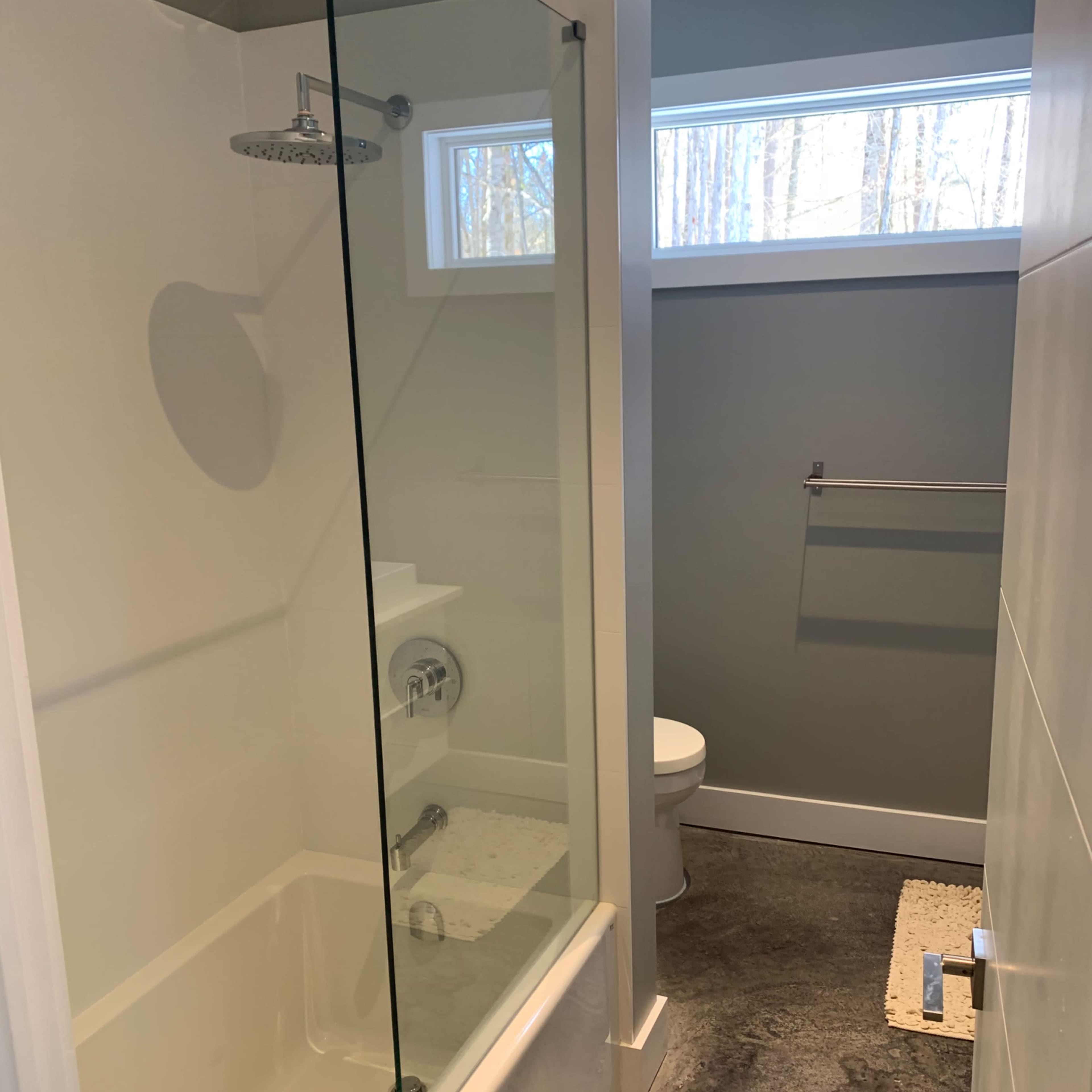 A modern bathroom featuring a glass shower, a bathtub, a toilet, and a towel rack against a light gray wall.
