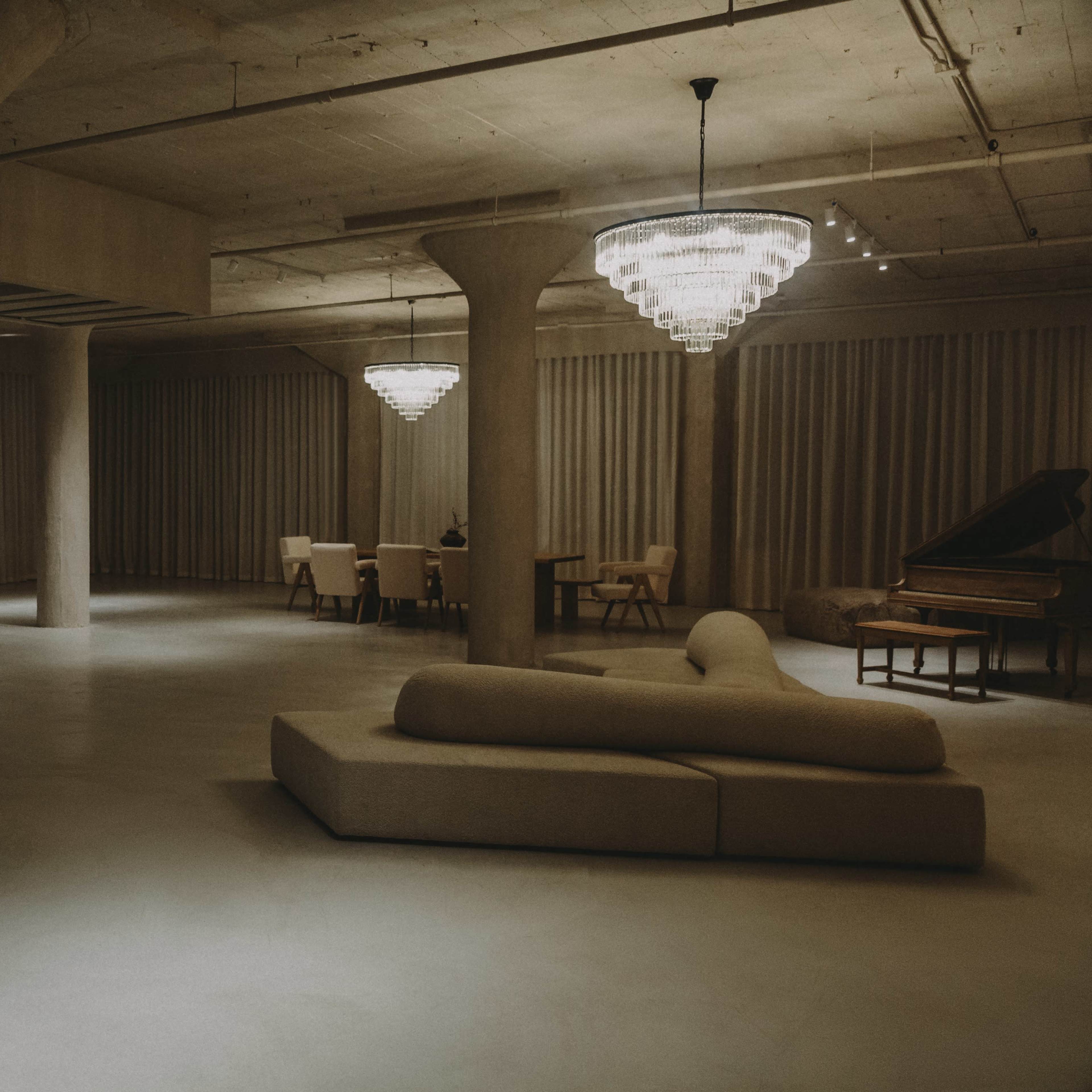 A spacious, dimly lit room with modern furniture, including a sectional sofa, dining area, and grand piano, surrounded by textured walls and soft drapery.