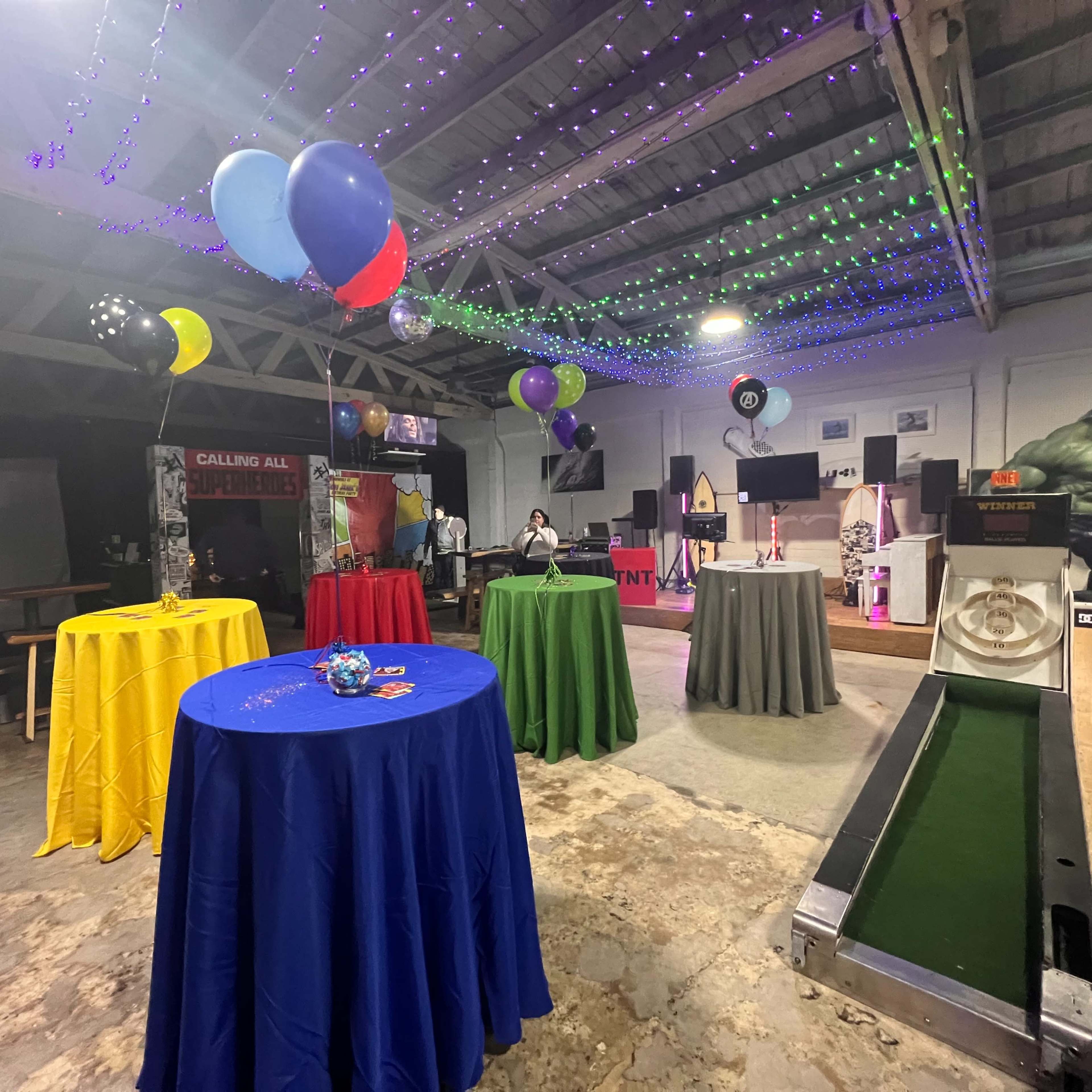 The scene shows a festive indoor space decorated with colorful tables, balloons, and string lights, featuring a mini-golf setup in the foreground.