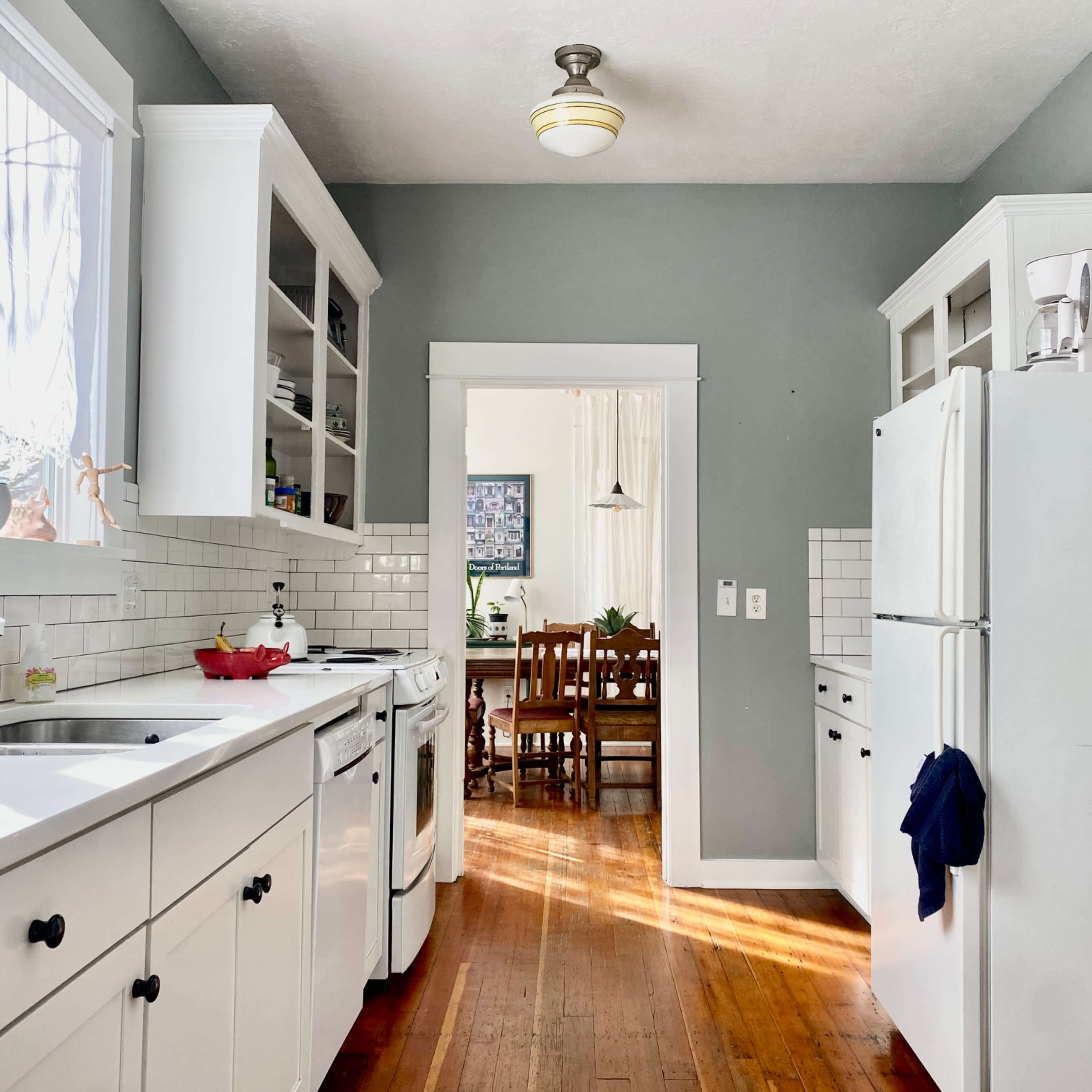 A narrow kitchen with white cabinetry, a refrigerator, and a view of an adjacent dining area through an open doorway.