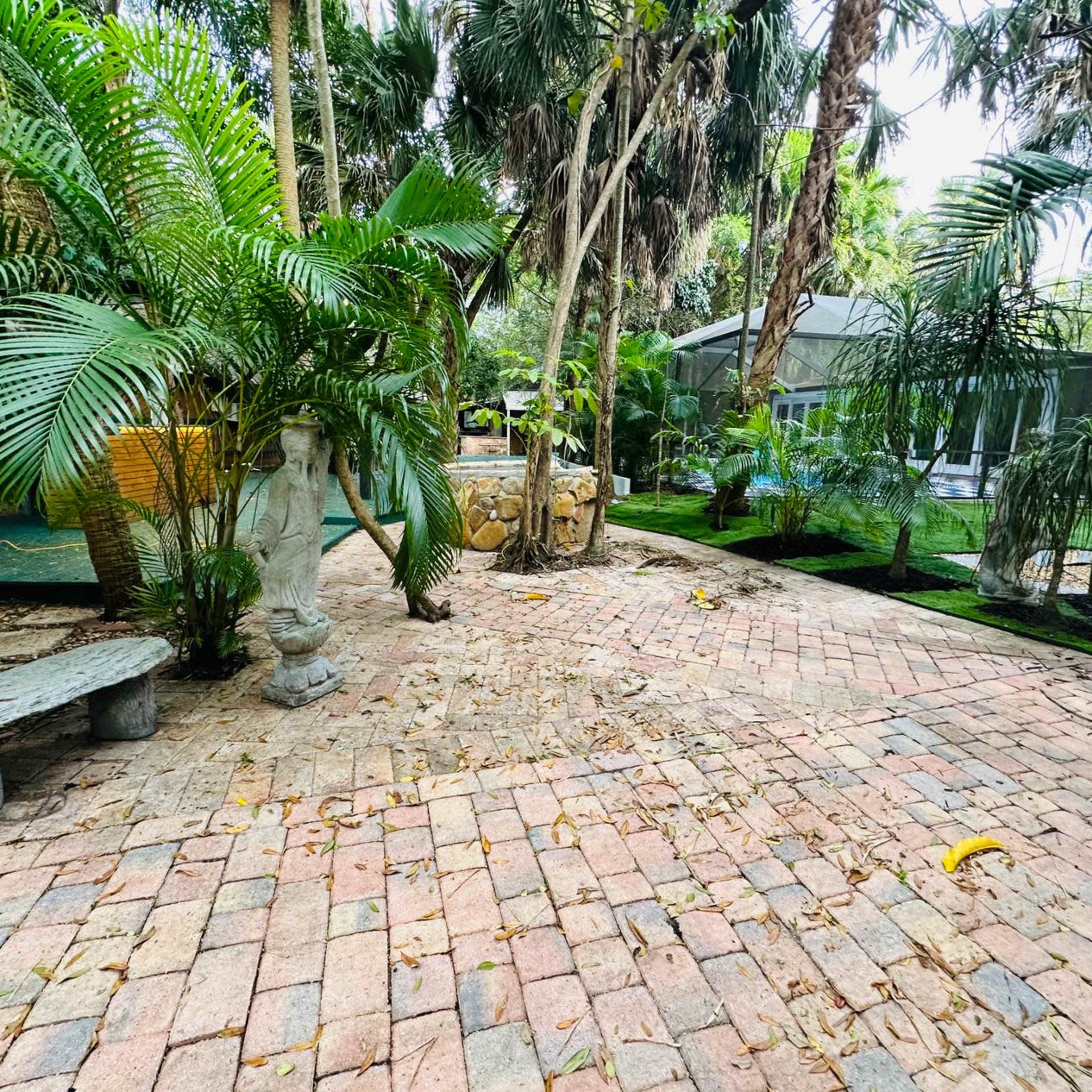 A landscaped garden area with brick pathways, stone benches, palm trees, and a small stone feature.