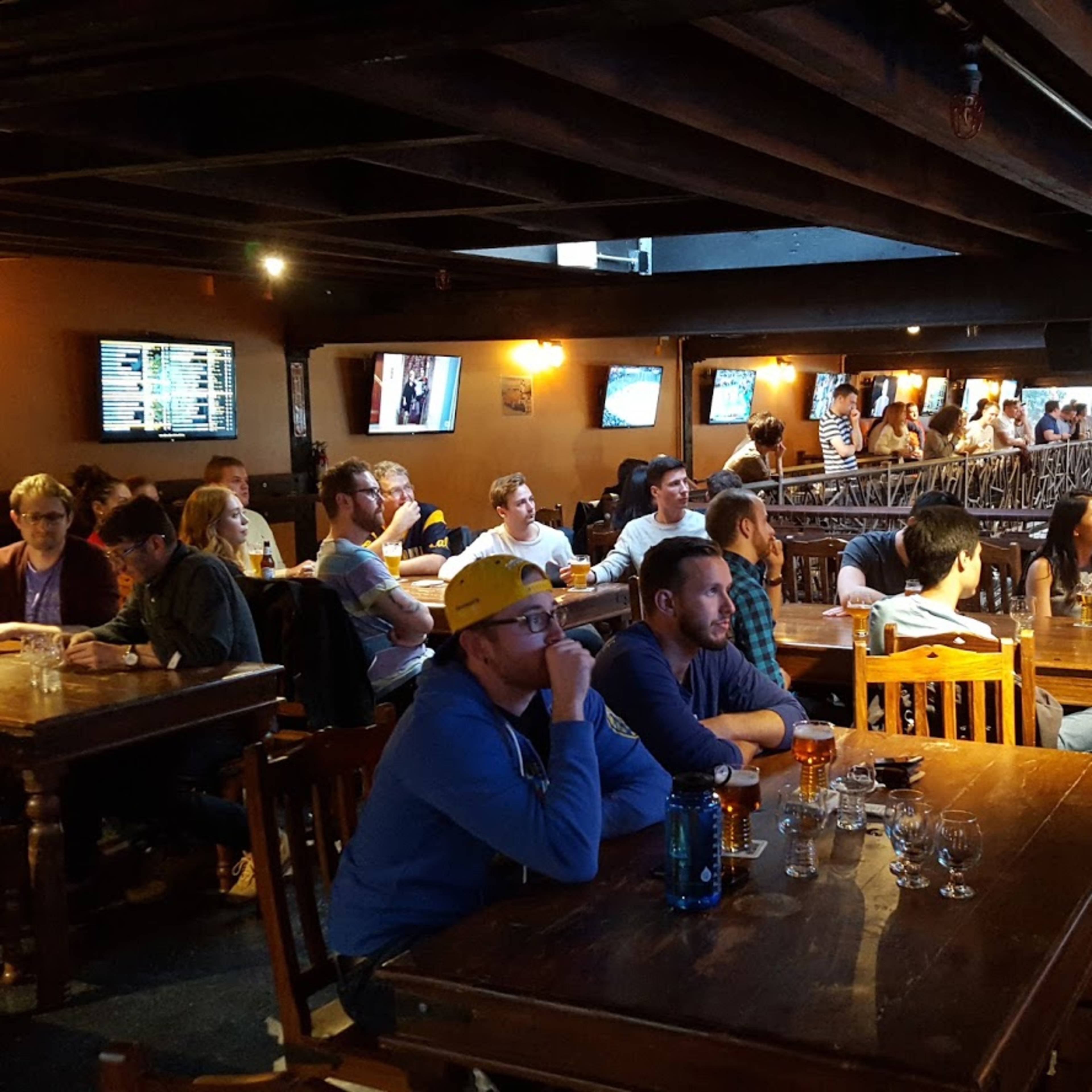 A large group of patrons sits at wooden tables inside a bar, watching screens displaying sports events.