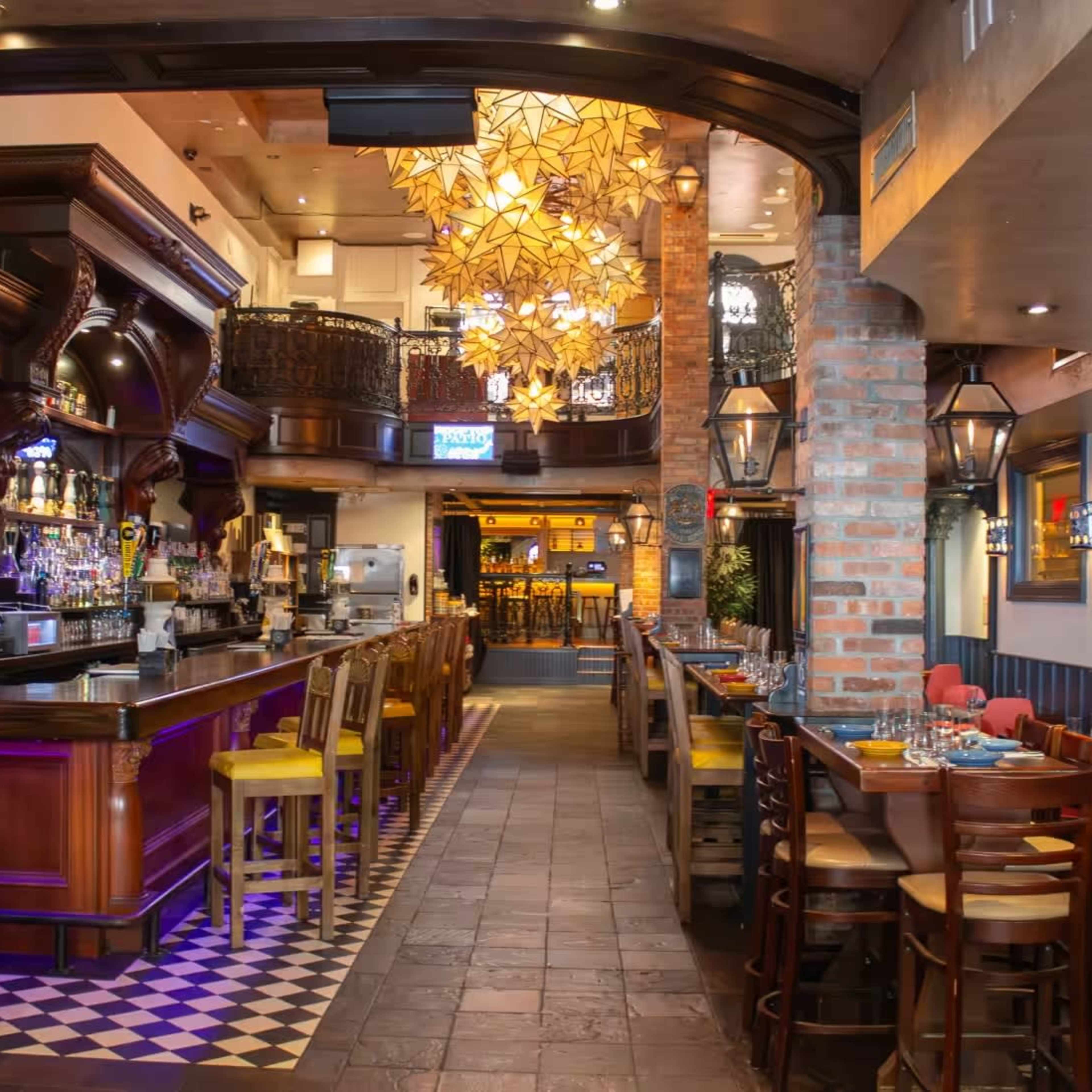 The interior of a restaurant featuring a bar area, colorful lighting fixtures, and dining tables arranged along a long, narrow space with decorative elements.